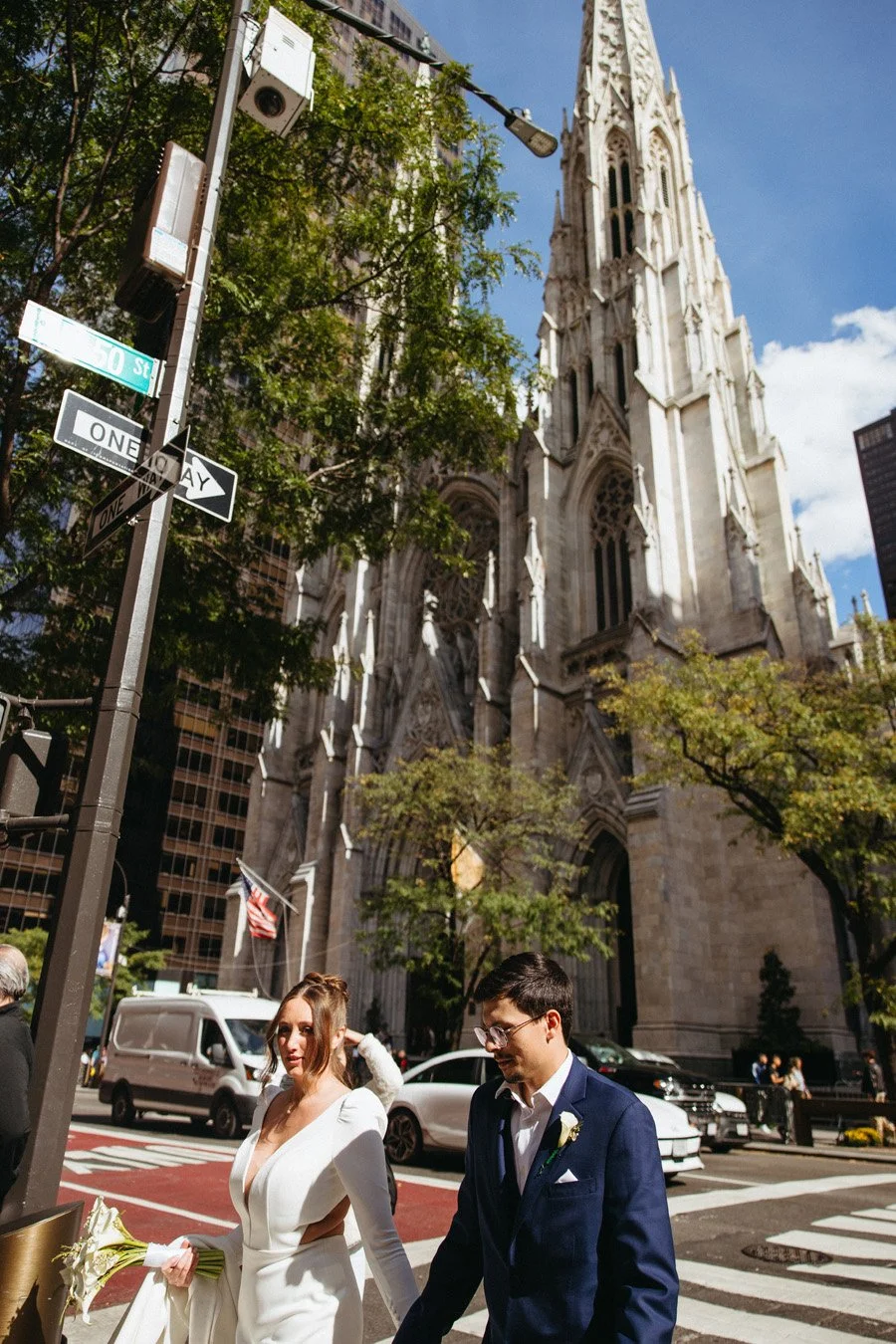 A bride and groom walking across a busy city street in front of a large Gothic-style church with a tall spire, trees, and traffic around them.