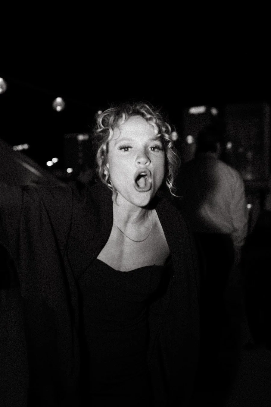 A woman with curly hair making a loud vocal expression, possibly shouting, in a dimly lit indoor setting.