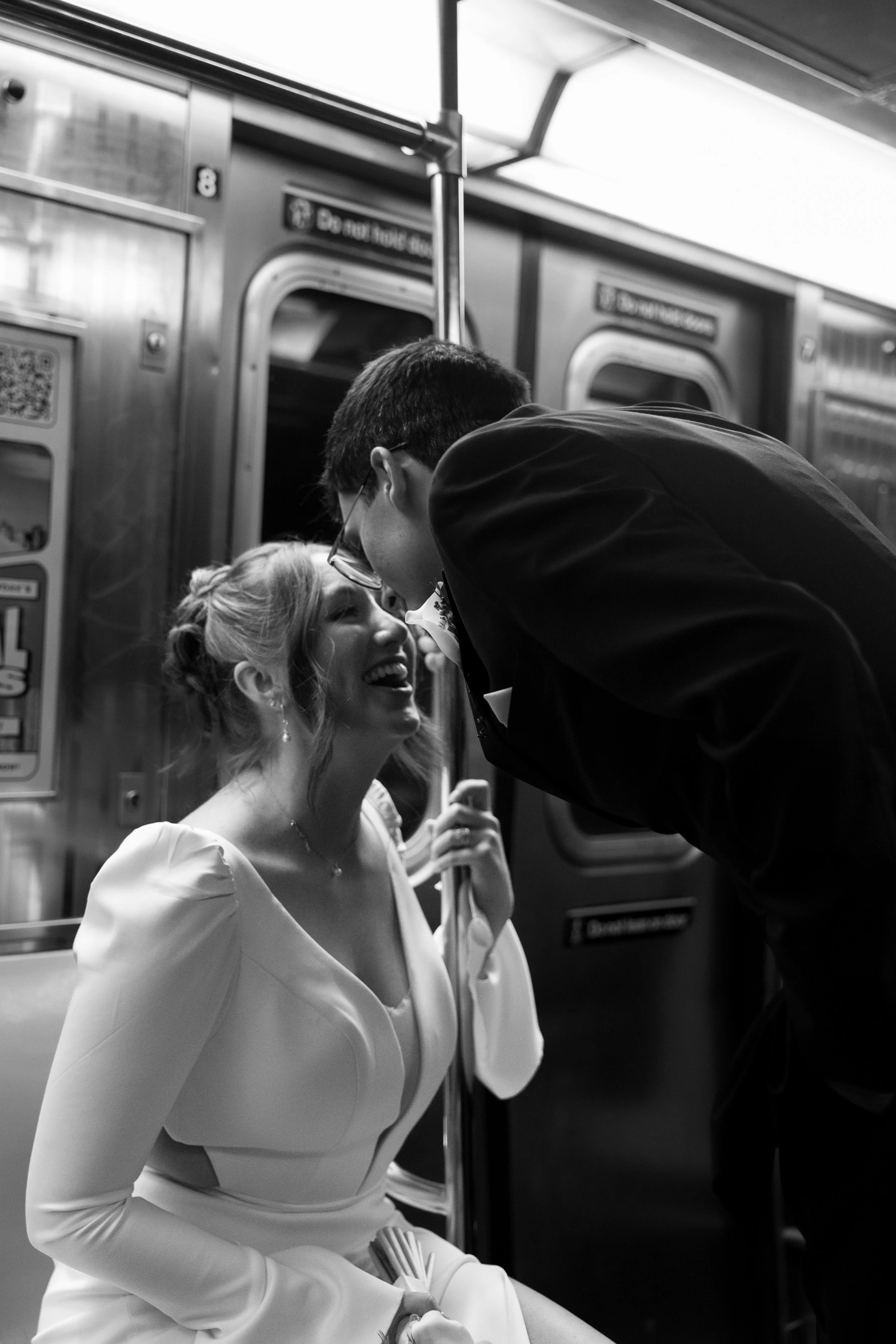 A black and white photo of a woman in a wedding dress smiling and laughing, leaning back slightly as a man in a suit leans in close to her inside a subway train.