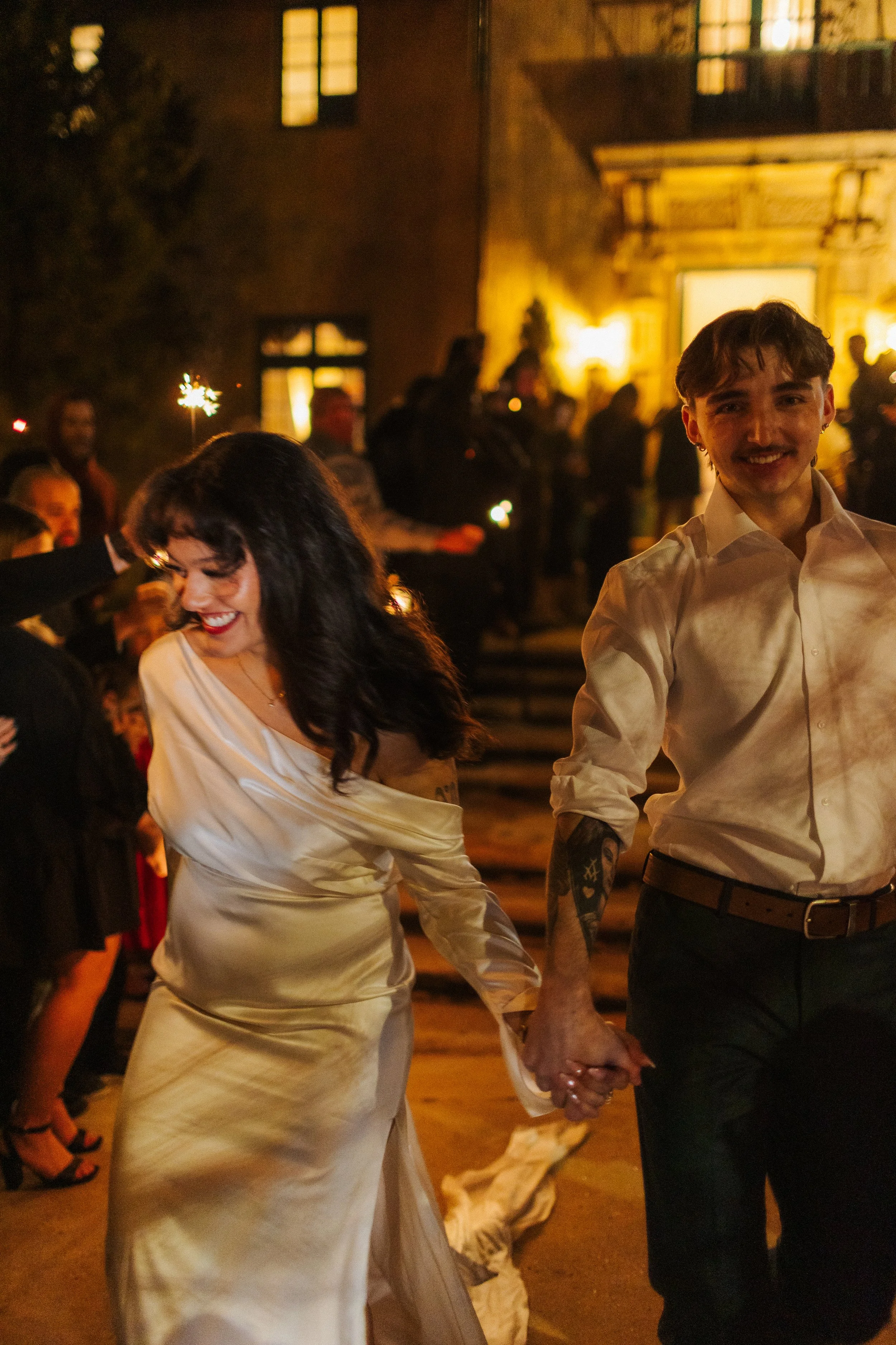 Couple holding hands and dancing during a wedding celebration at night, surrounded by guests with sparklers outside a lit building.