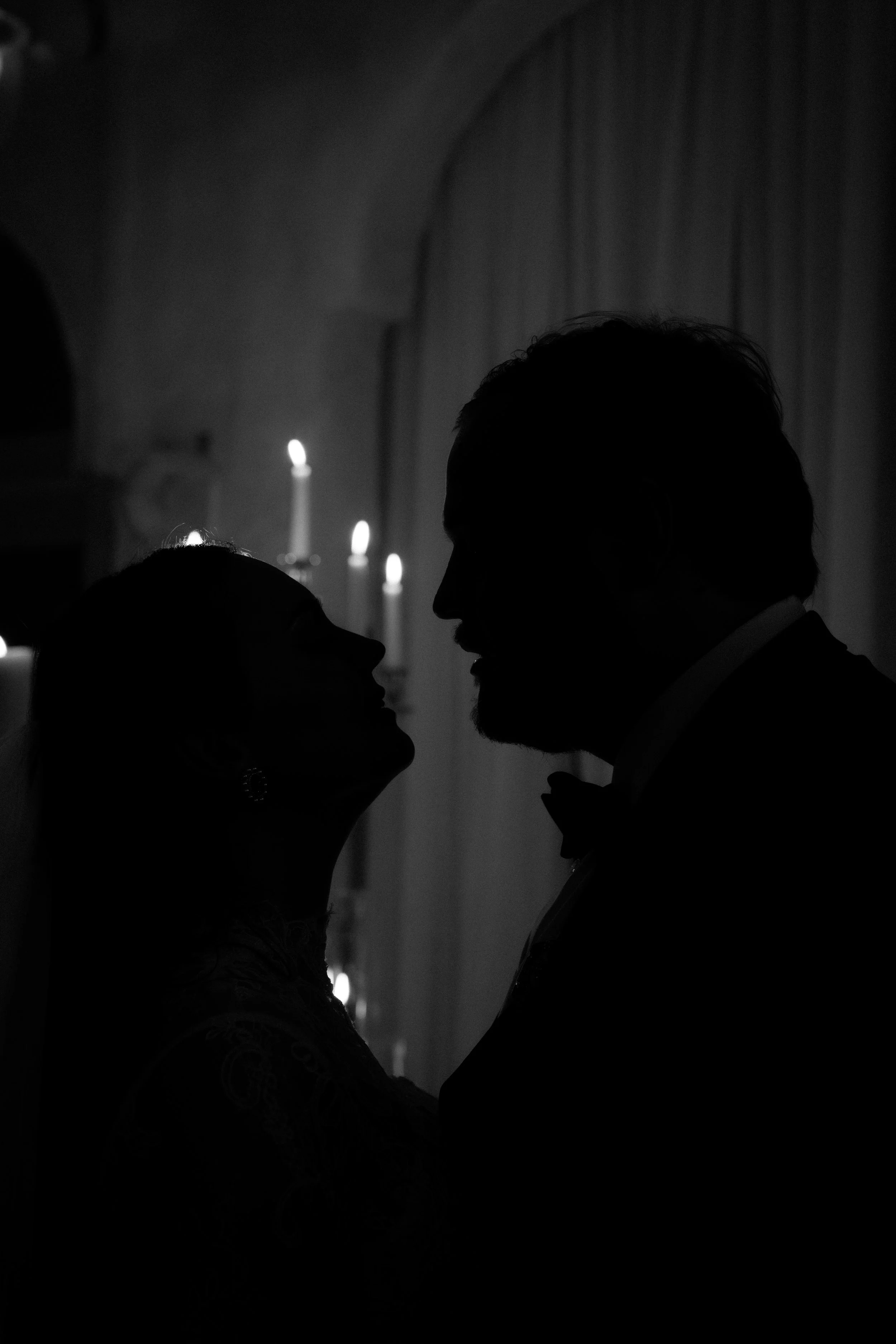 A black and white image of the silhouette of a man and woman going in for a kiss, backlit by candles.