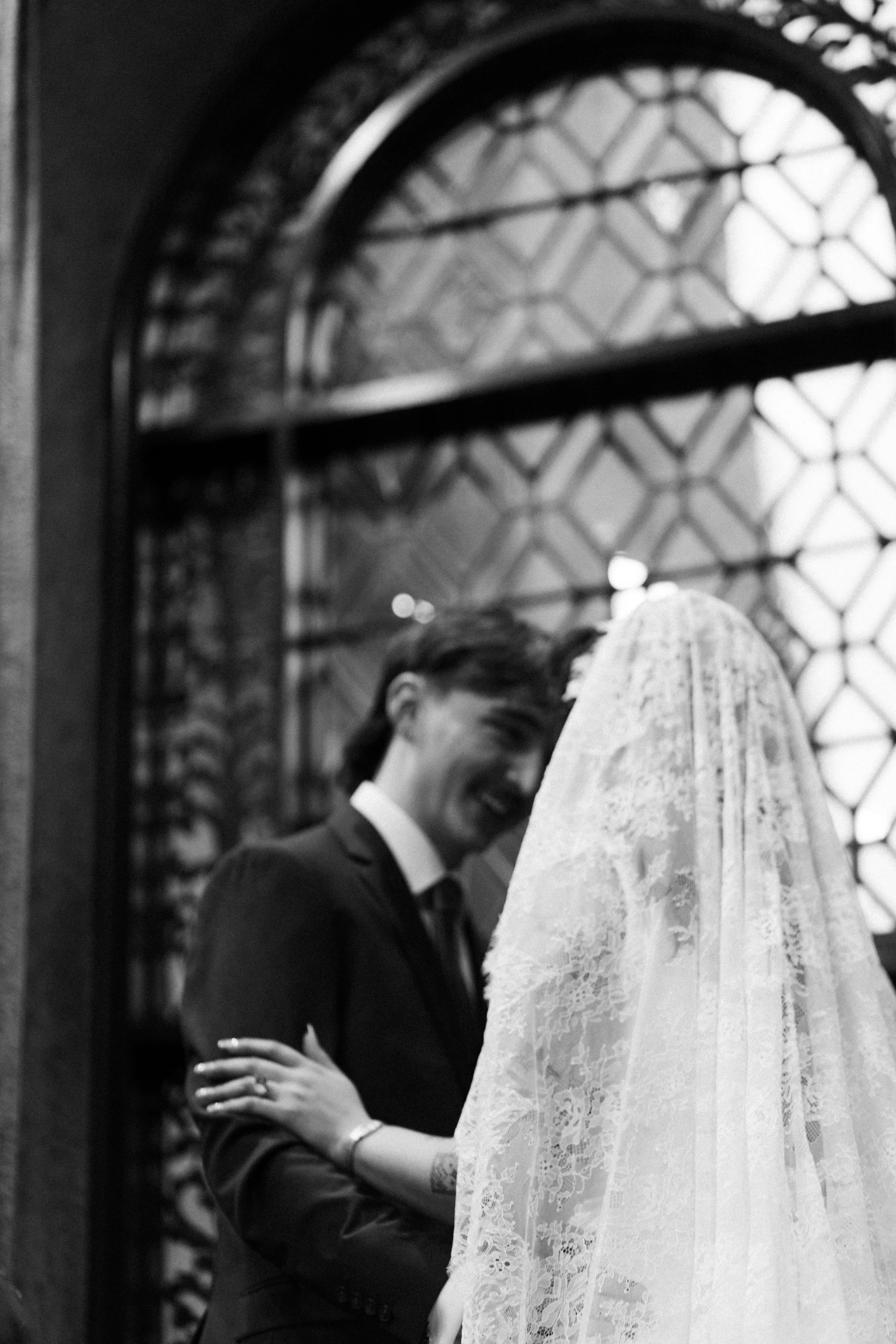 A bride and groom face each other in a close-up black and white photograph, smiling and holding hands, in front of a decorative window.