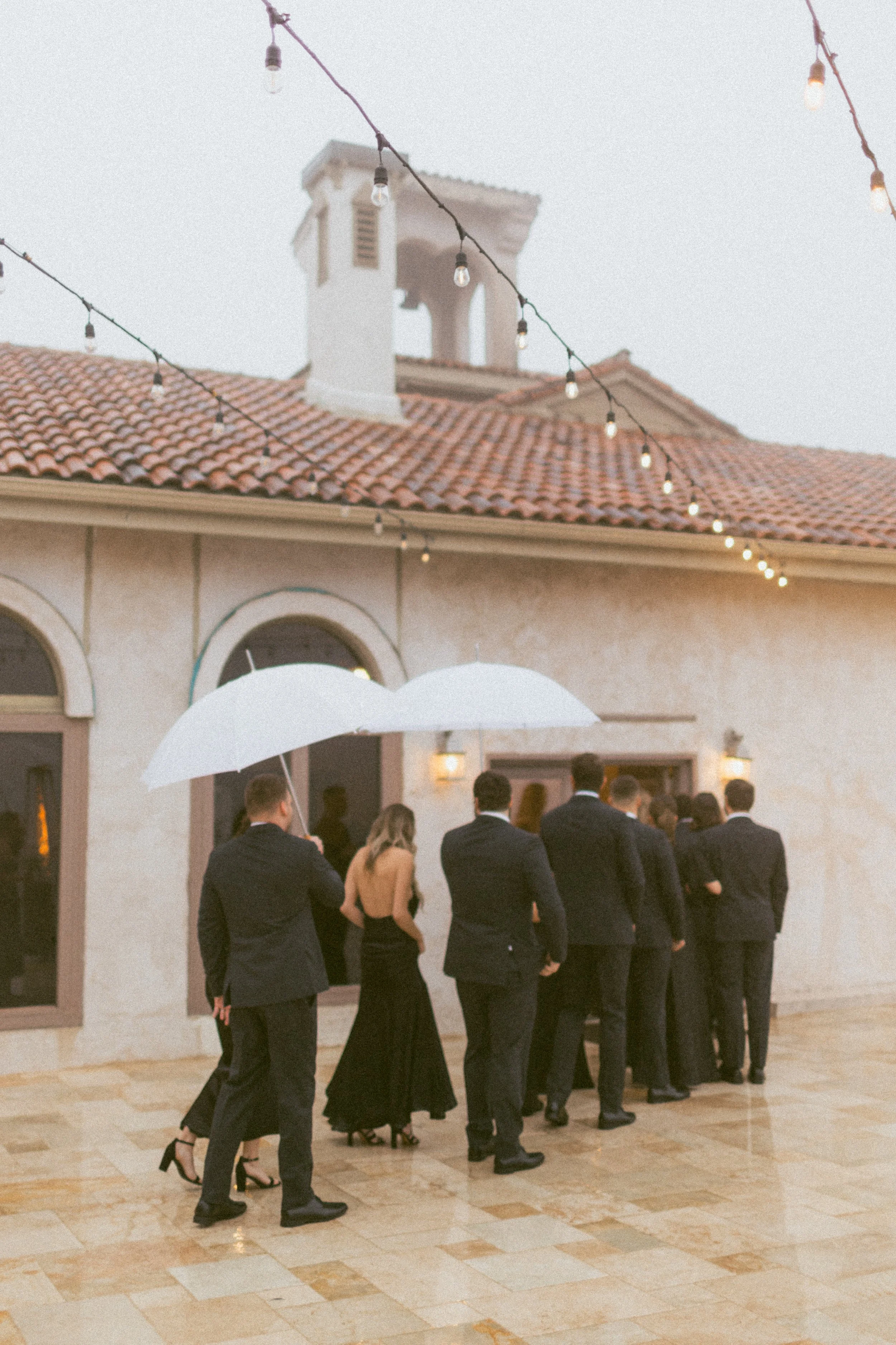 Group of people in formal attire waiting in line under umbrellas outside a building with string lights overhead.