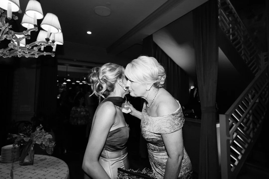 Two women are whispering to each other at a formal event in a dimly lit room with a chandelier and a staircase in the background.
