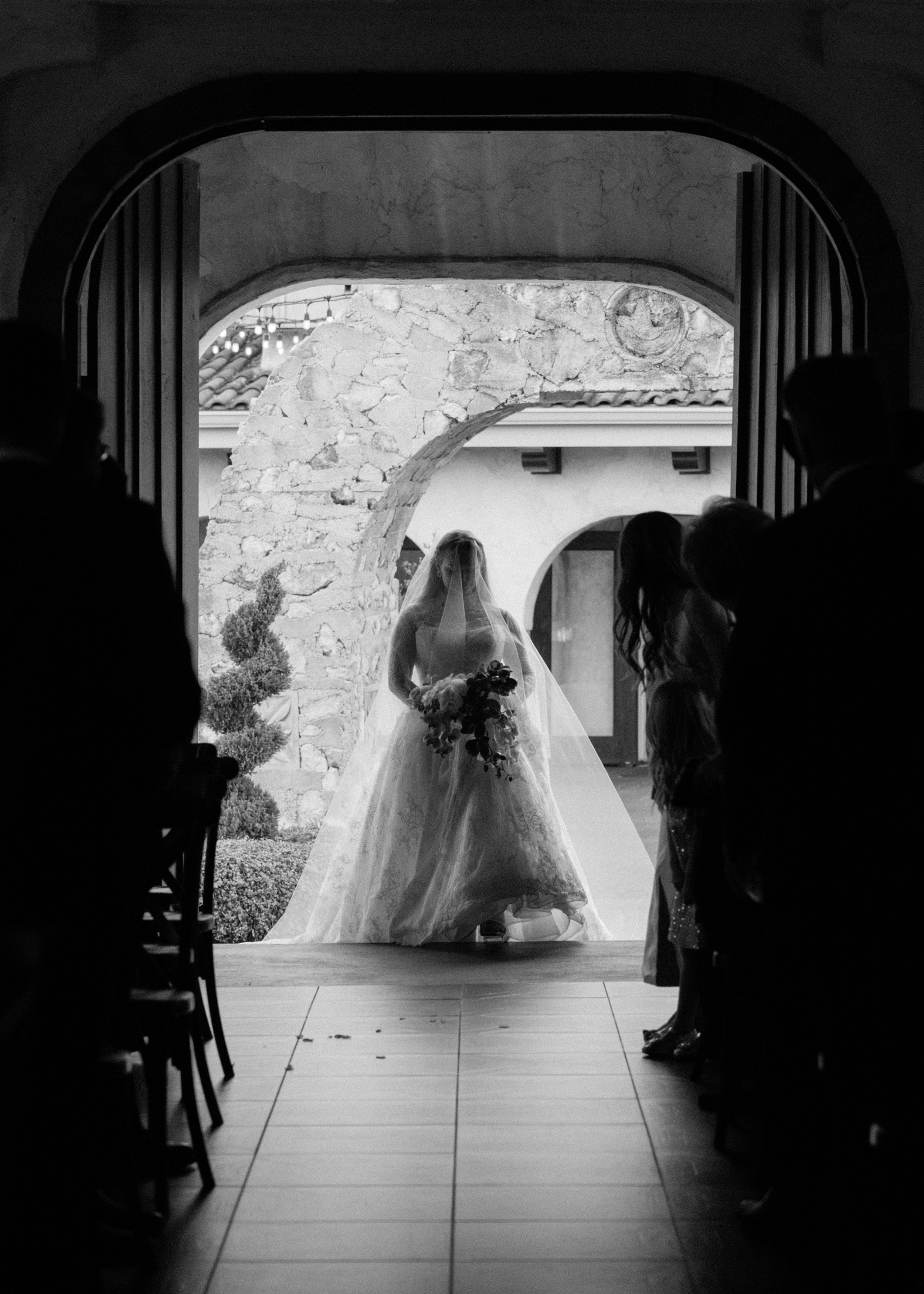 A black and white image of a woman in a wedding dress with a long veil over her face, entering a chapel.