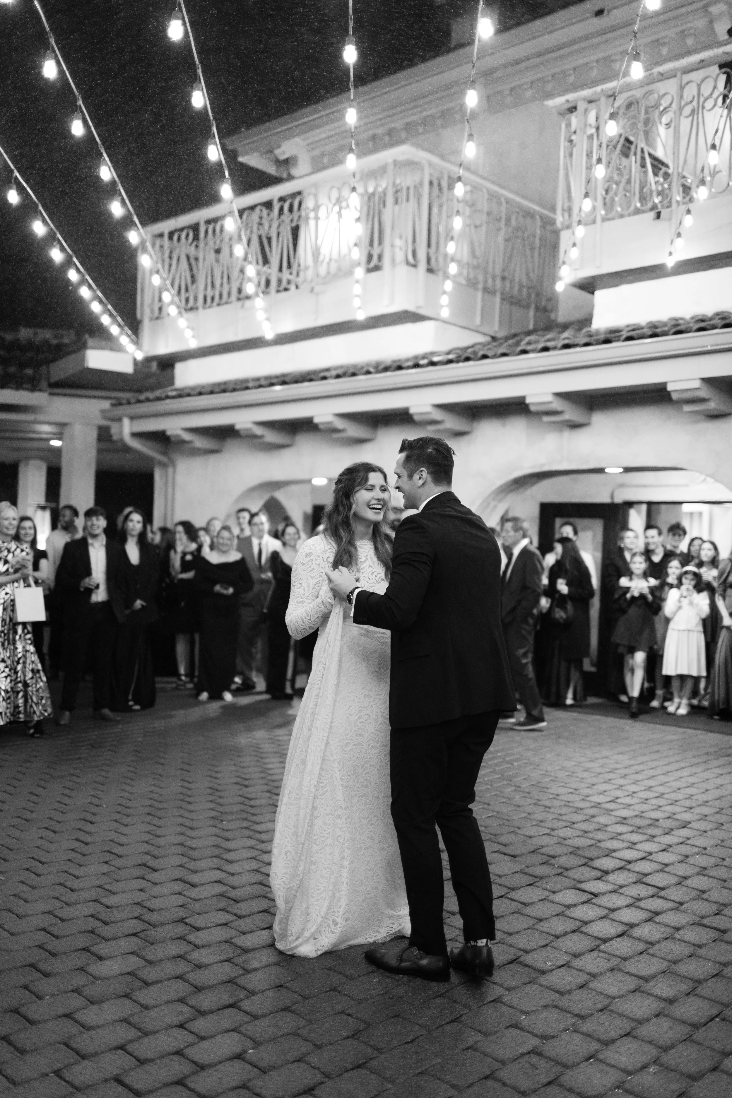 A couple is dancing at their wedding reception, smiling and looking at each other, surrounded by guests in an outdoor courtyard at night with string lights overhead.
