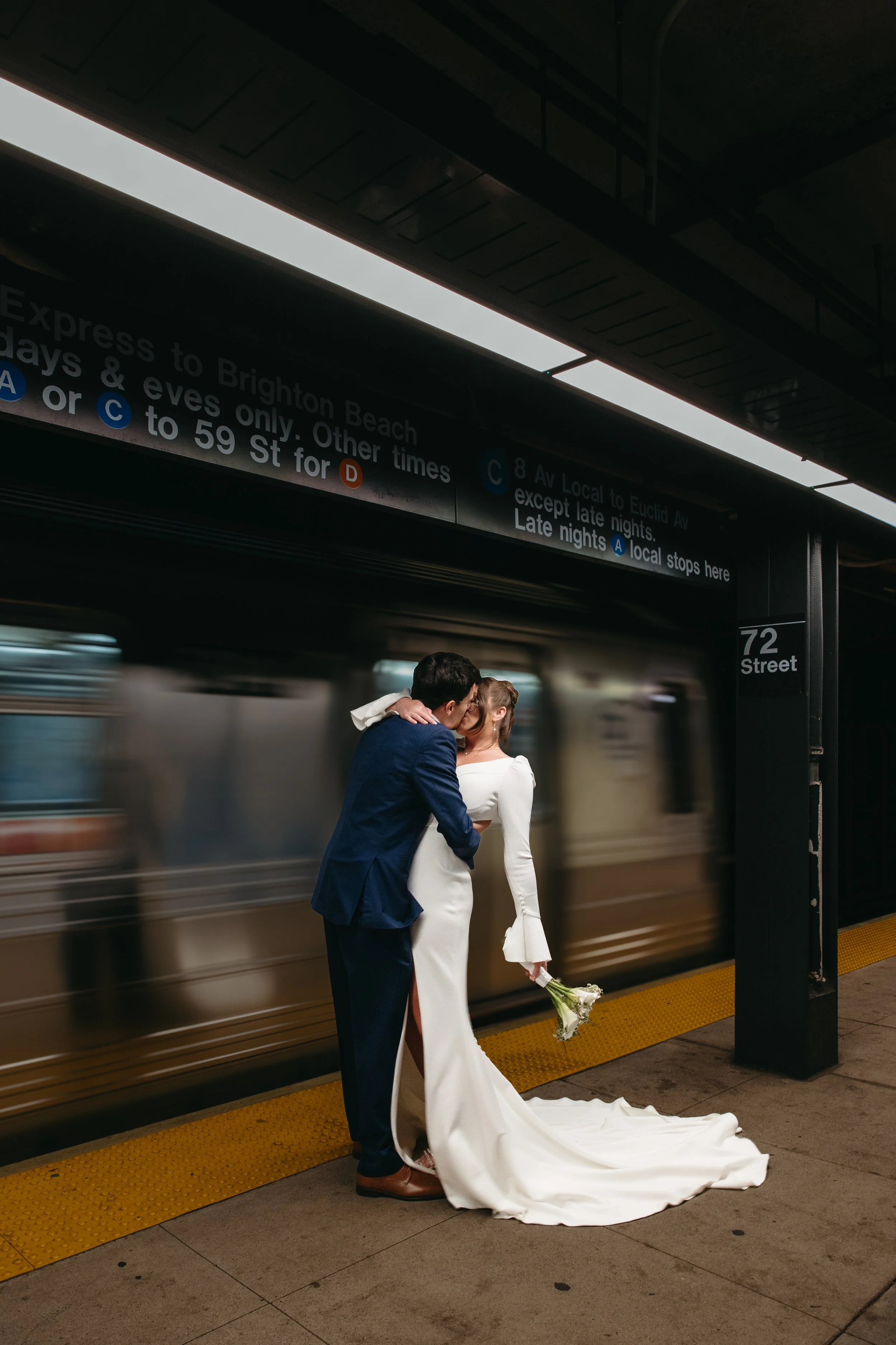 A couple in wedding attire sharing a kiss on a subway platform, with a moving train in the background.