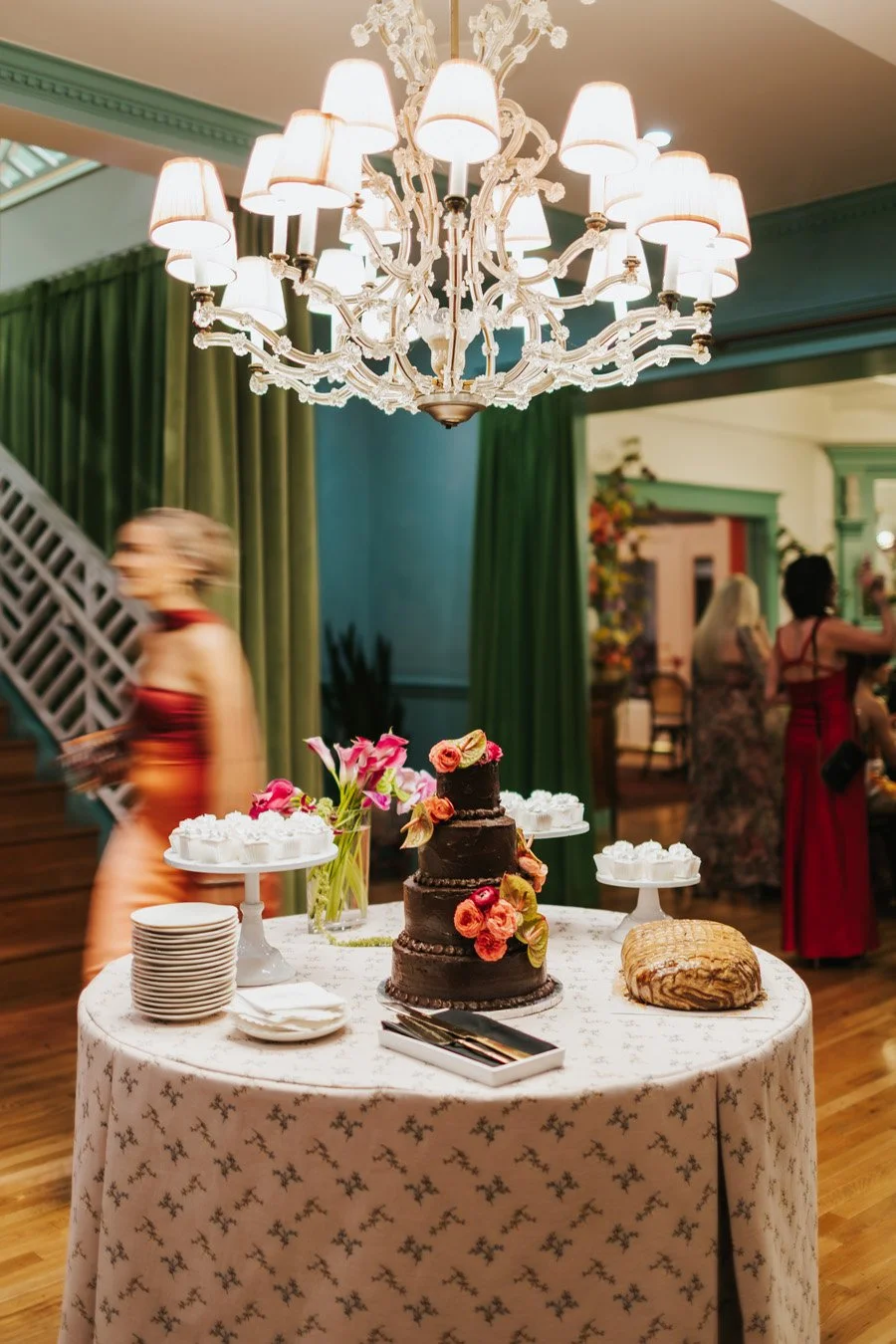 Decorative three-tier chocolate cake with orange and pink flowers on a table with white plates and cups at a celebration, with blurred guests in the background and a chandelier overhead.
