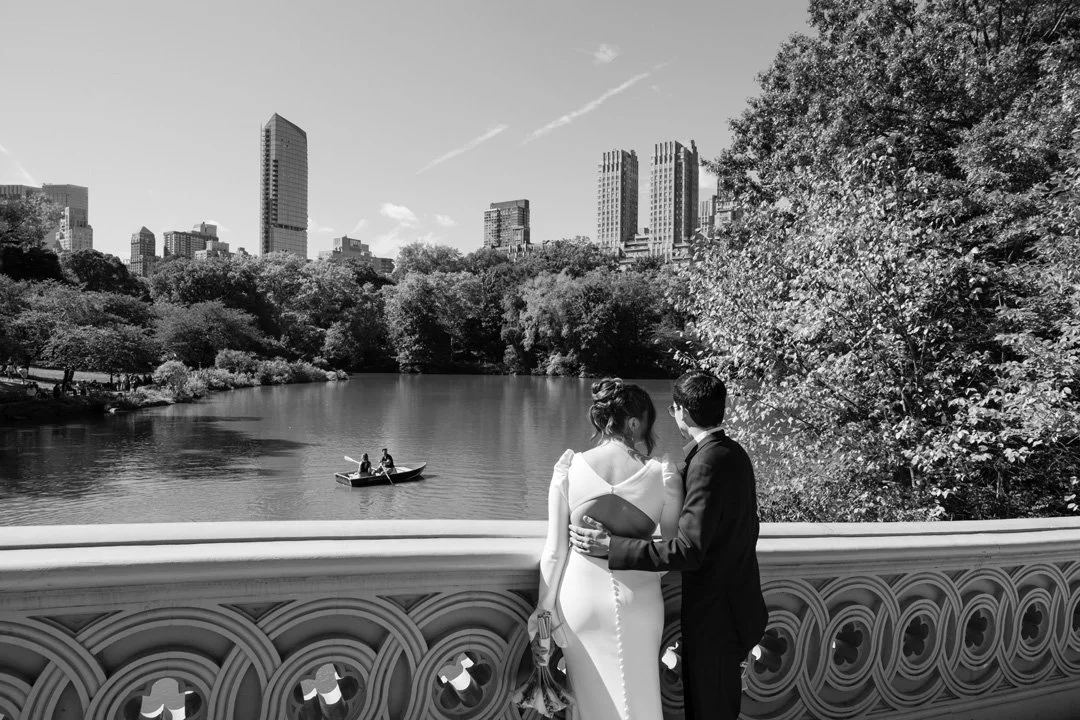 A black-and-white photo of a couple standing on a bridge overlooking a lake in Central Park, New York City. The woman wears a white dress, and the man wears a dark suit. They are holding each other and looking at the lake, with a city skyline in the background and trees surrounding the lake.