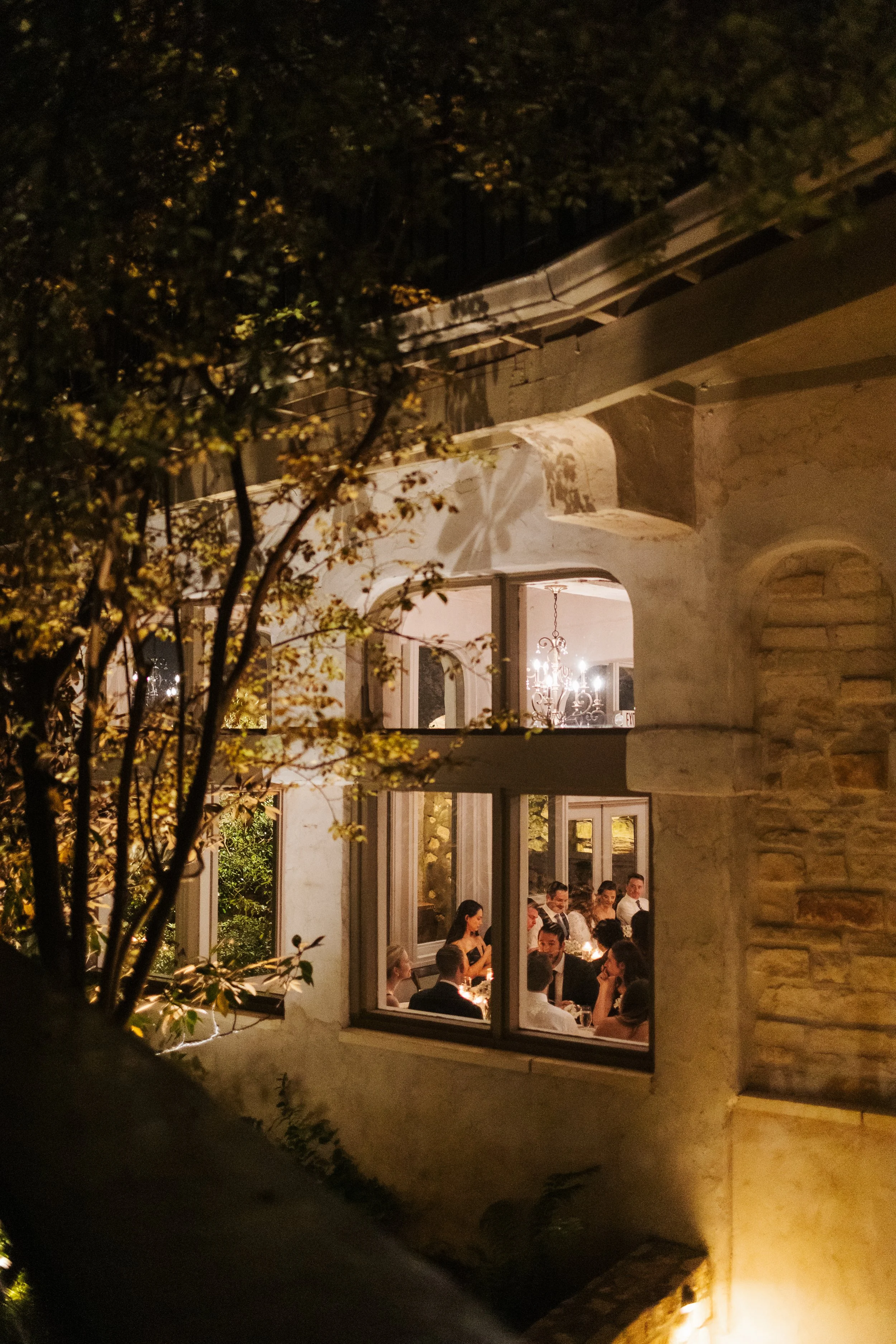 People dining inside a warmly lit restaurant during the night, viewed through a window from outside, with foliage partially obscuring the view.