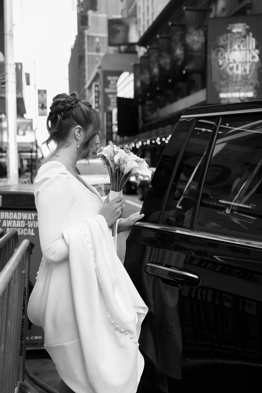 A woman dressed in a white gown holding a bouquet of flowers, standing beside a parked vehicle on a city street.