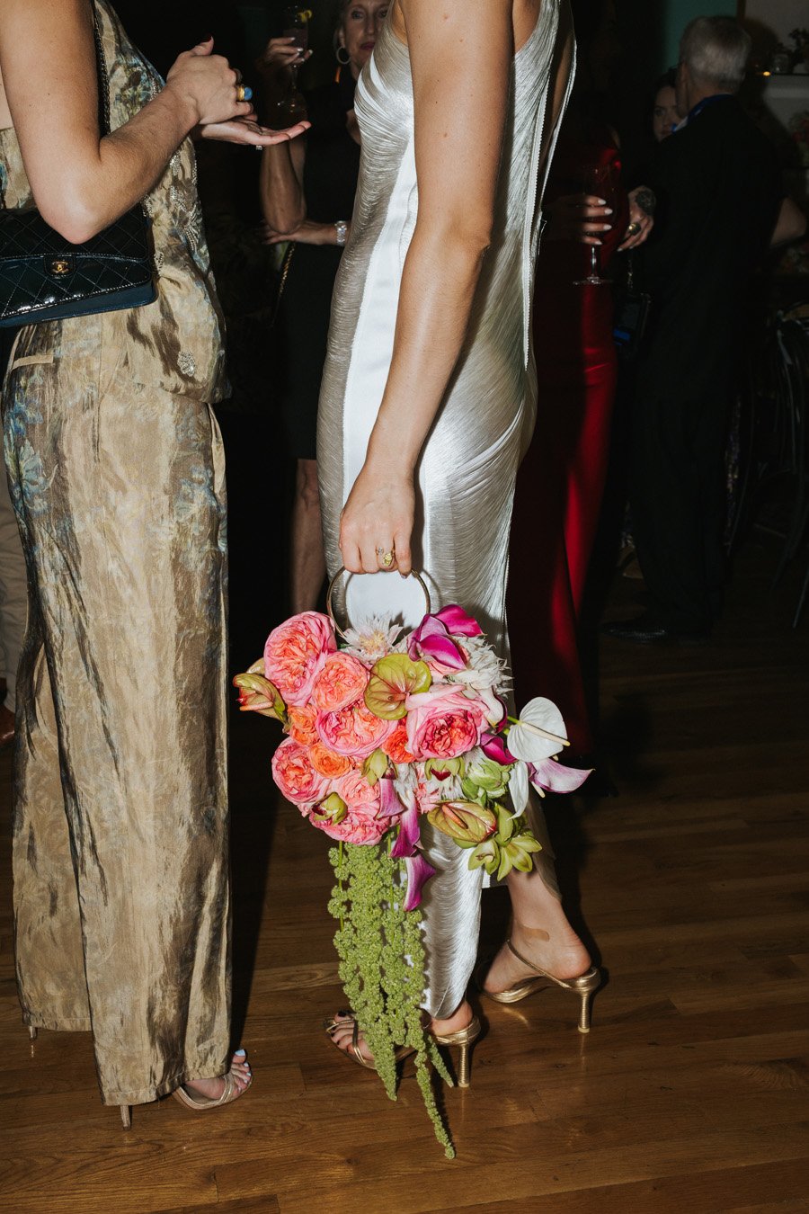 A woman in a silk wedding dress holding a floral handbag with pink roses, purple orchids, and greenery at a social event.