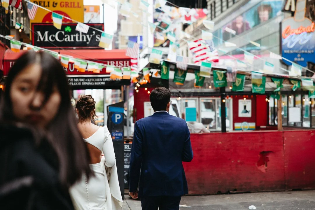 A man in a blue suit walking through a busy street with Irish and American flags and pub signs, with blurry women in the foreground.