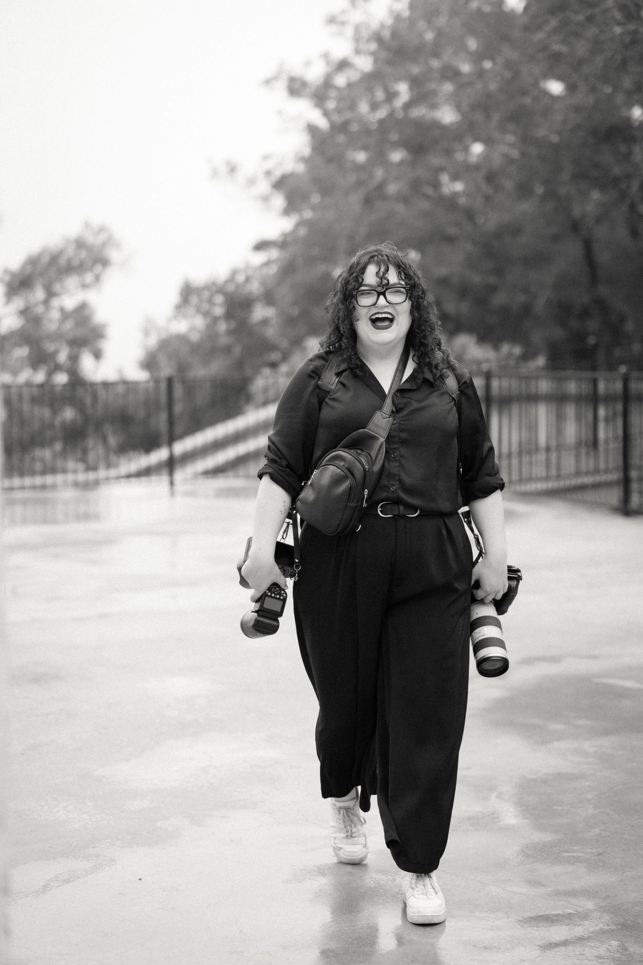 A woman with curly hair and glasses, smiling, walking outdoors with a camera in one hand and a smaller camera or phone in the other. She is wearing a dark shirt, long pants, sneakers, and carrying multiple bags, with trees and a fence in the background.