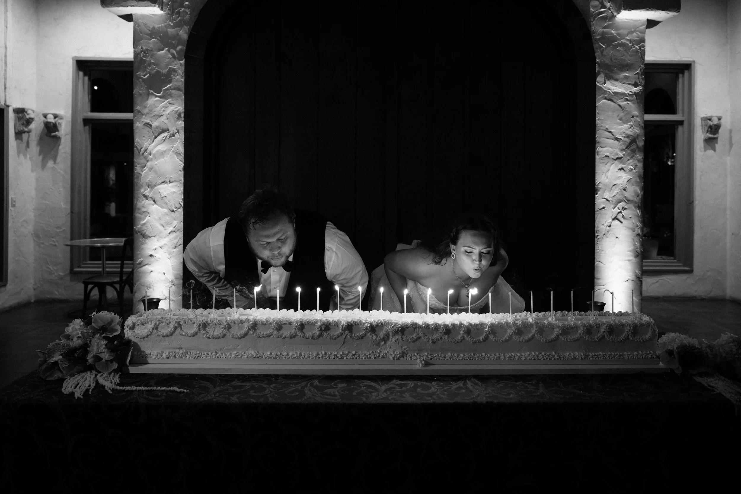 A black and white image of a man and woman in wedding attire leaning over to blow out candles on a long wedding cake.
