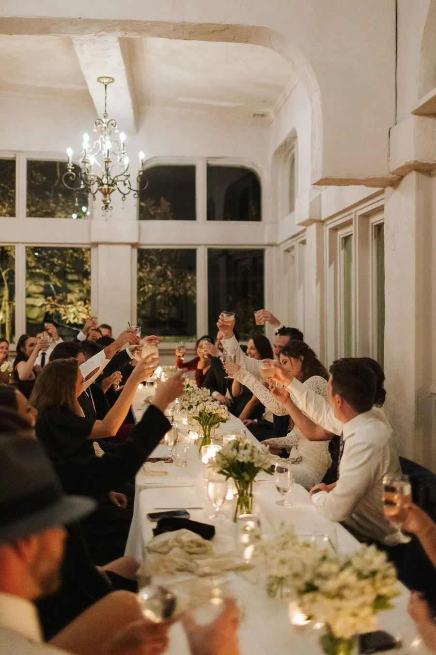 A wedding reception with guests seated at a long decorated table, raising glasses in a toast, inside a bright room with large windows and a chandelier.