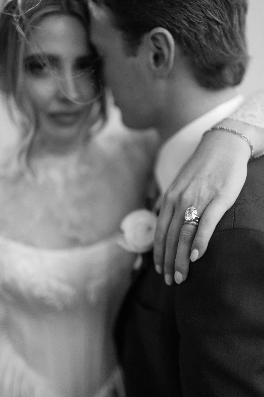Black and white close-up of a couple, with the woman’s hand resting on the man’s shoulder, showing a large engagement ring and wedding band.
