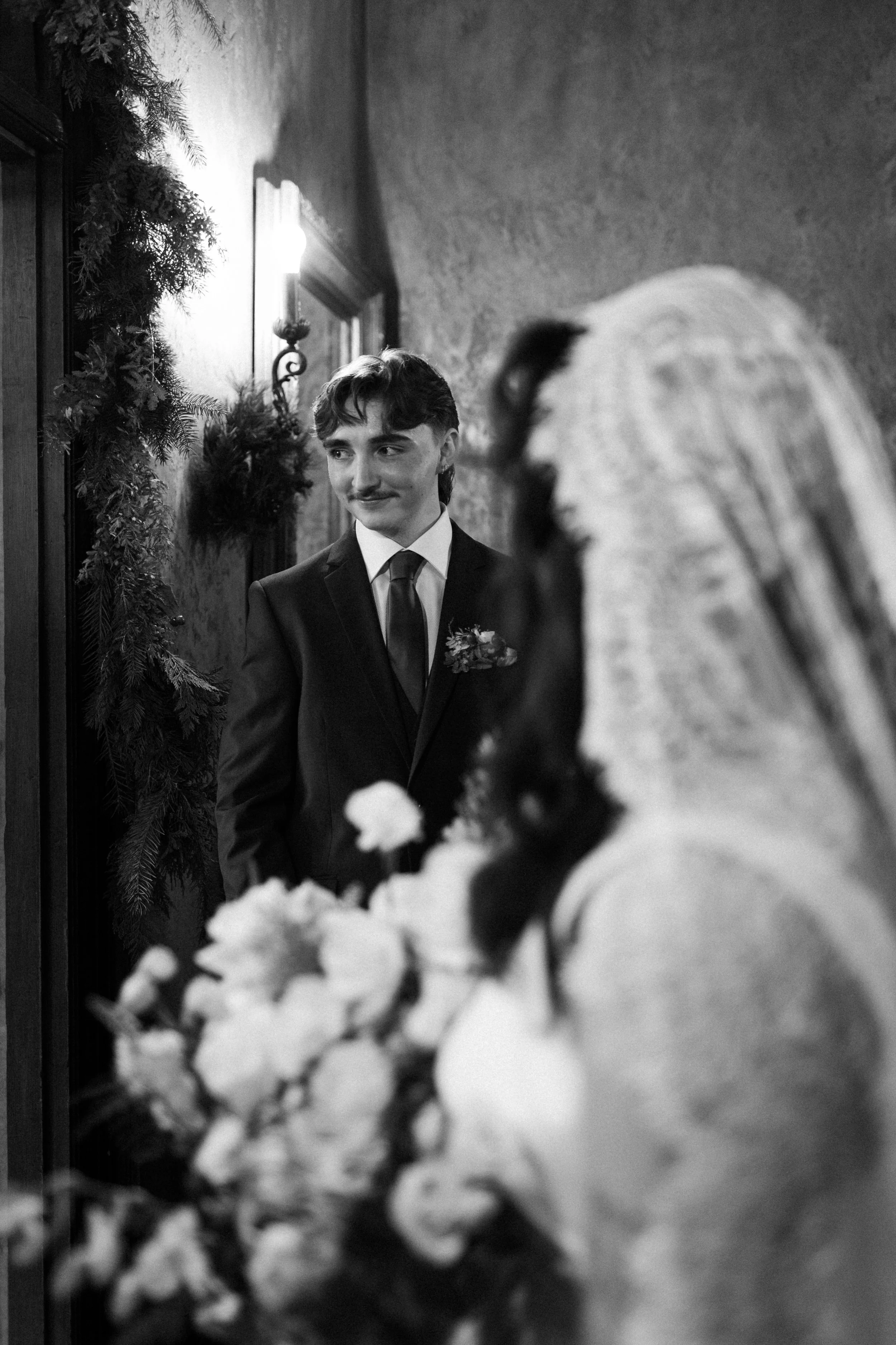 A groom in a dark suit and tie looking at a bride on their wedding day, with floral bouquet in the foreground, in a warmly decorated indoor setting.