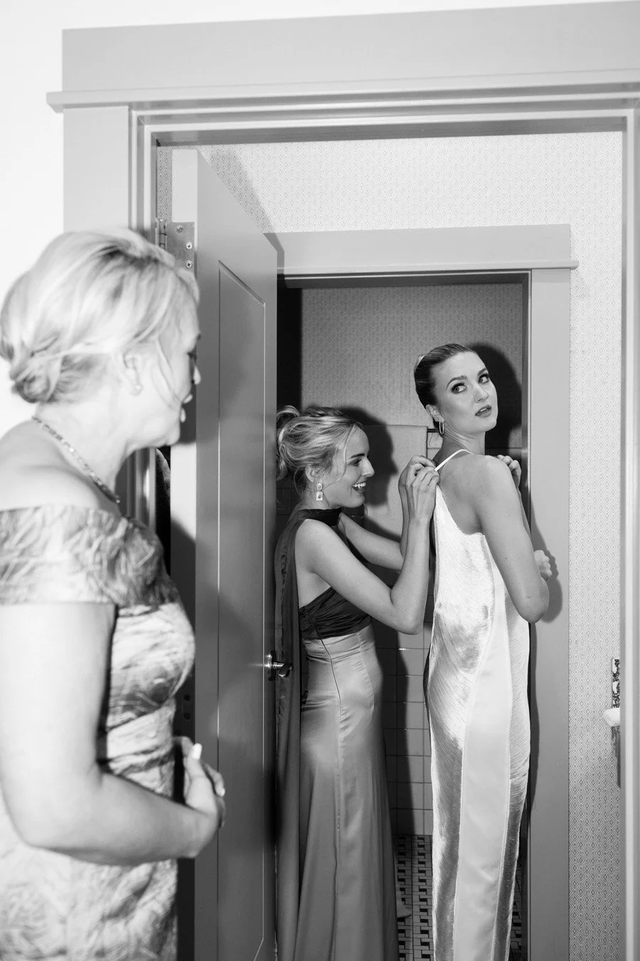 A woman helps another woman adjust her dress in a fitting room, with a third woman observing.