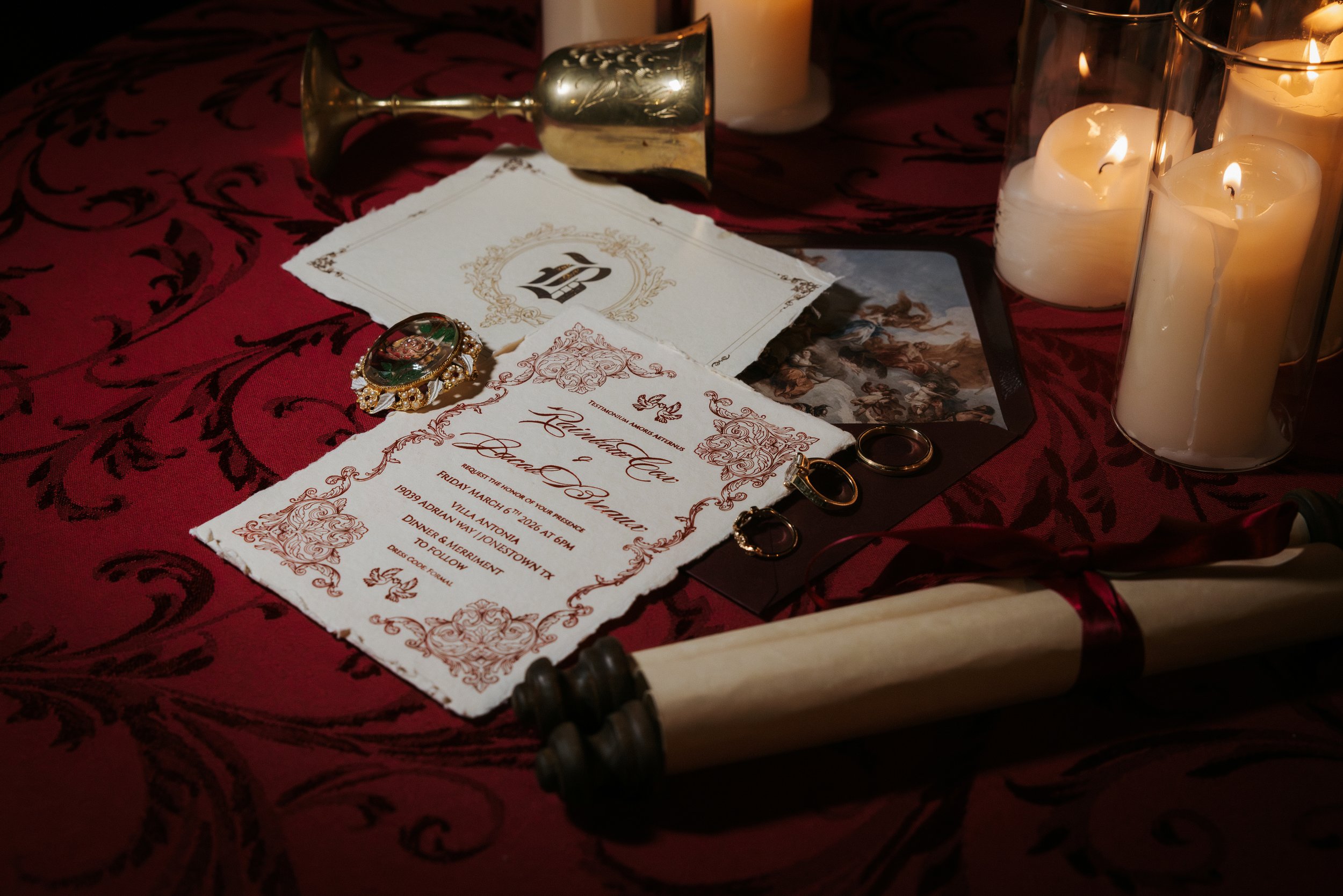 A flat lay of a wedding invite suite with candles, a scroll, and wedding rings, with a red background.