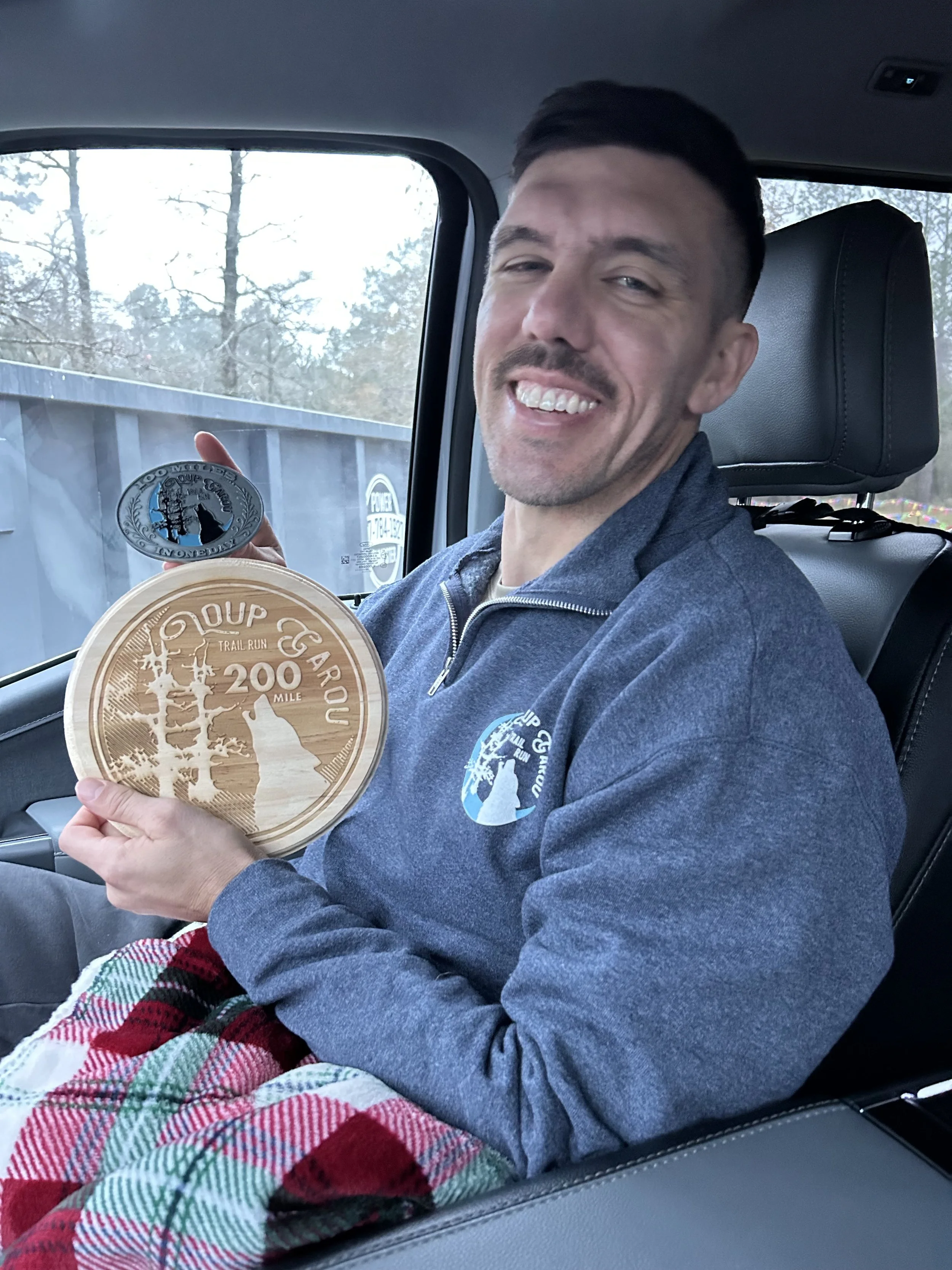 A smiling man with short dark hair and a beard sitting in a car, holding a wooden medal with the inscription '200 MILE TRAIL RUN'. He is wearing a dark blue jacket and a plaid blanket on his lap, with trees visible outside the car window.