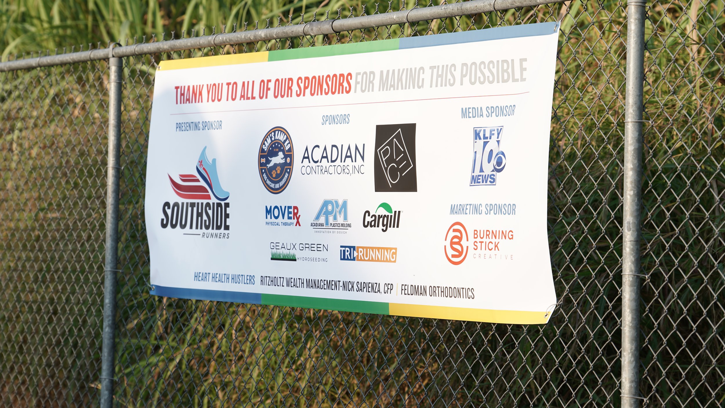 A large banner on a chain link fence thanking sponsors of an event. Logos of sponsors including Southside Runners, Acadian Contractors, Inc., PAC, KLFY News, MOVE Physical Therapy, AP Plastics Molding, Cargill, Heart Health Hustlers, TRI Running, Geaux Green Hydroseeding, and others are displayed.