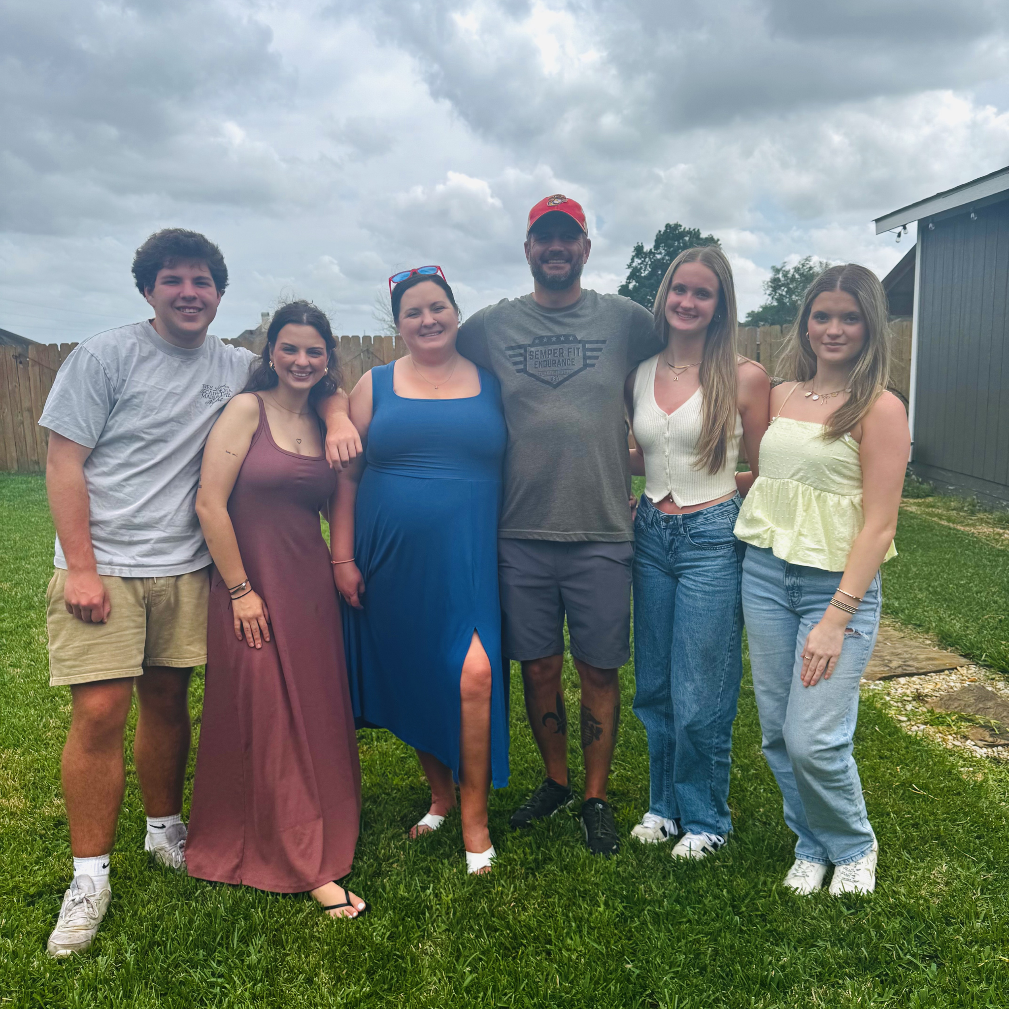 Group of six young adults, four women and two men, standing outdoors on grass with a cloudy sky in the background. They are smiling and posing for a photo.