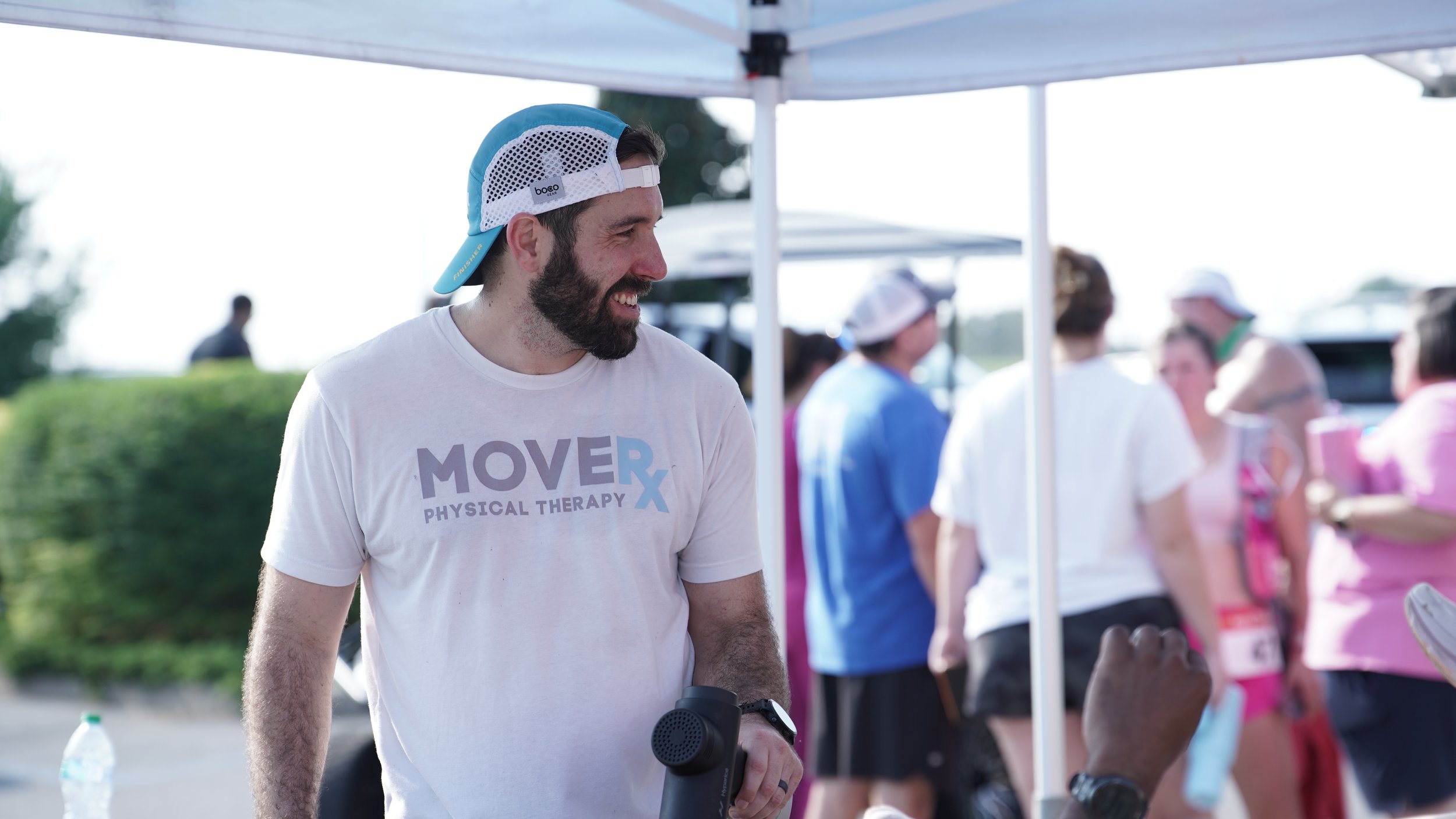 A man smiling at a race event wearing a white T-shirt, a baseball cap backward, and holding a water bottle, with other participants and a tent in the background.