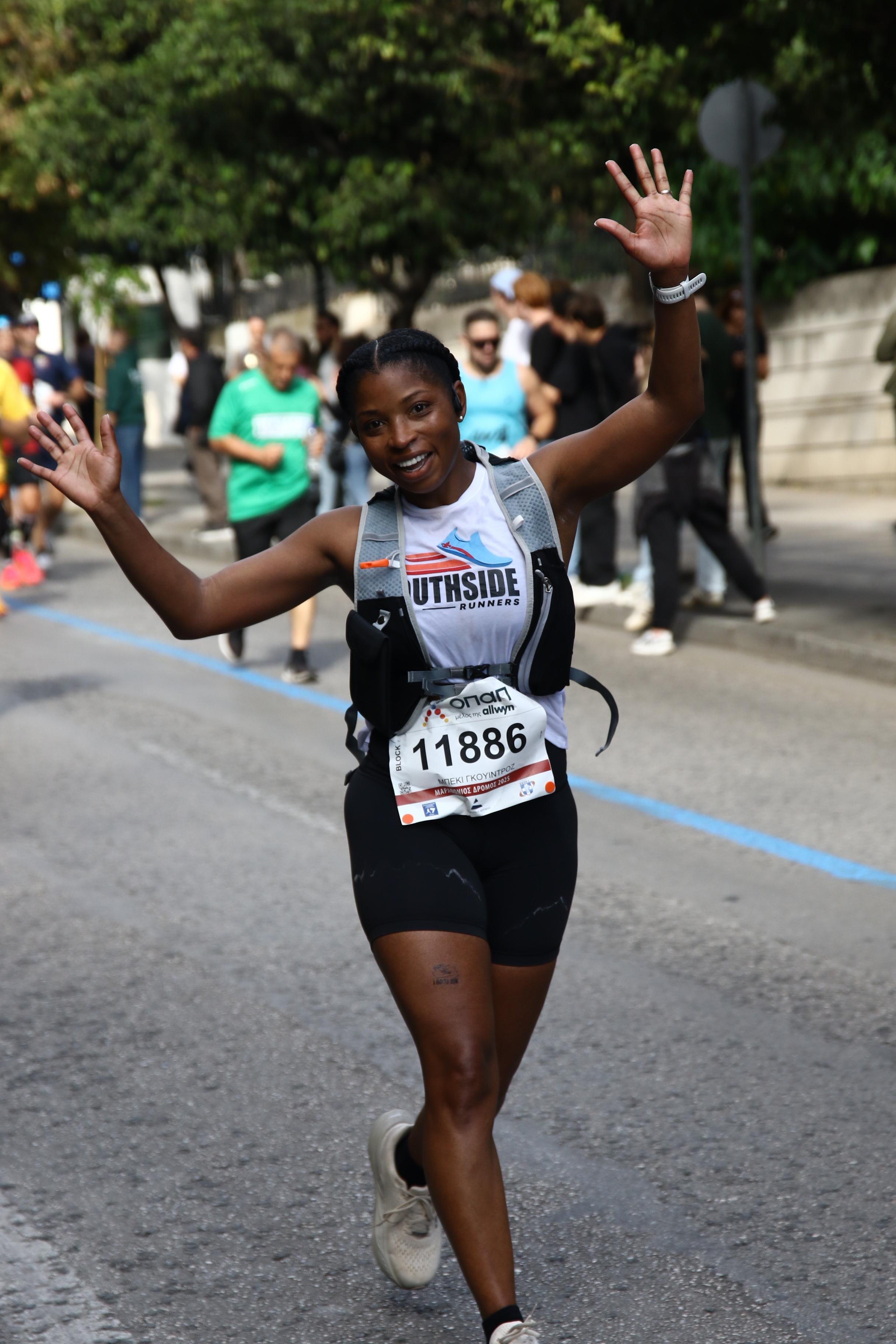 Young woman participating in a marathon, smiling, with her hands raised, wearing a race bib with the number 11886, running on a city street with other runners in the background and trees lining the street.