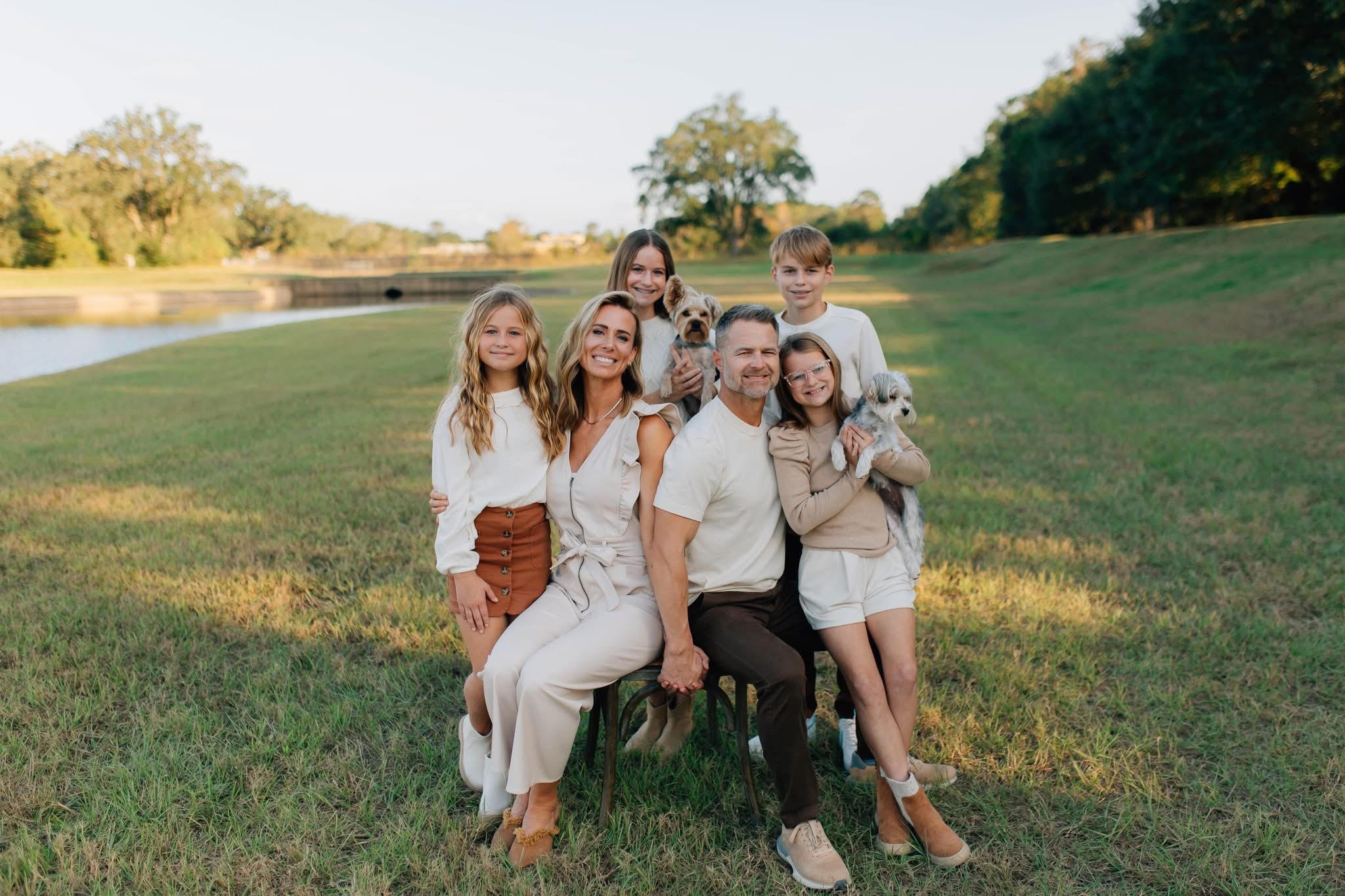 A family of six, including two children, two adults, and two dogs, poses for a photo outdoors in a grassy area near a pond, with trees and a blue sky in the background.