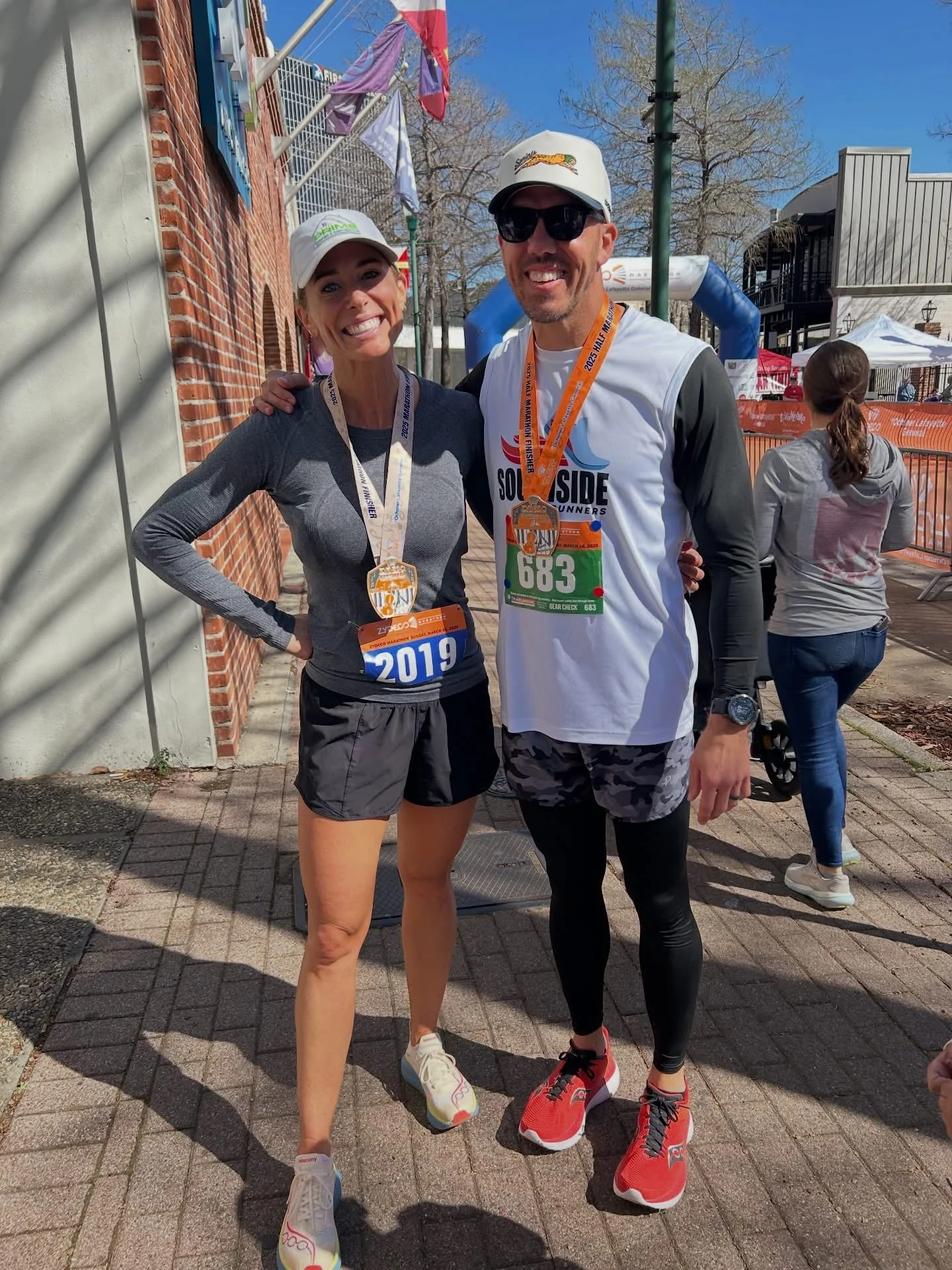 Two runners, a woman and a man, smiling at the camera after completing a race. They are wearing athletic gear and medals around their necks, with race bibs pinned to their shirts. The woman has a grey long-sleeve top, black shorts, white running shoes, and a white cap. The man has a white sleeveless shirt, black leggings, red running shoes, a white cap with a logo, and sunglasses. The background shows a brick building, some banners, and a few other people, with clear blue skies overhead.