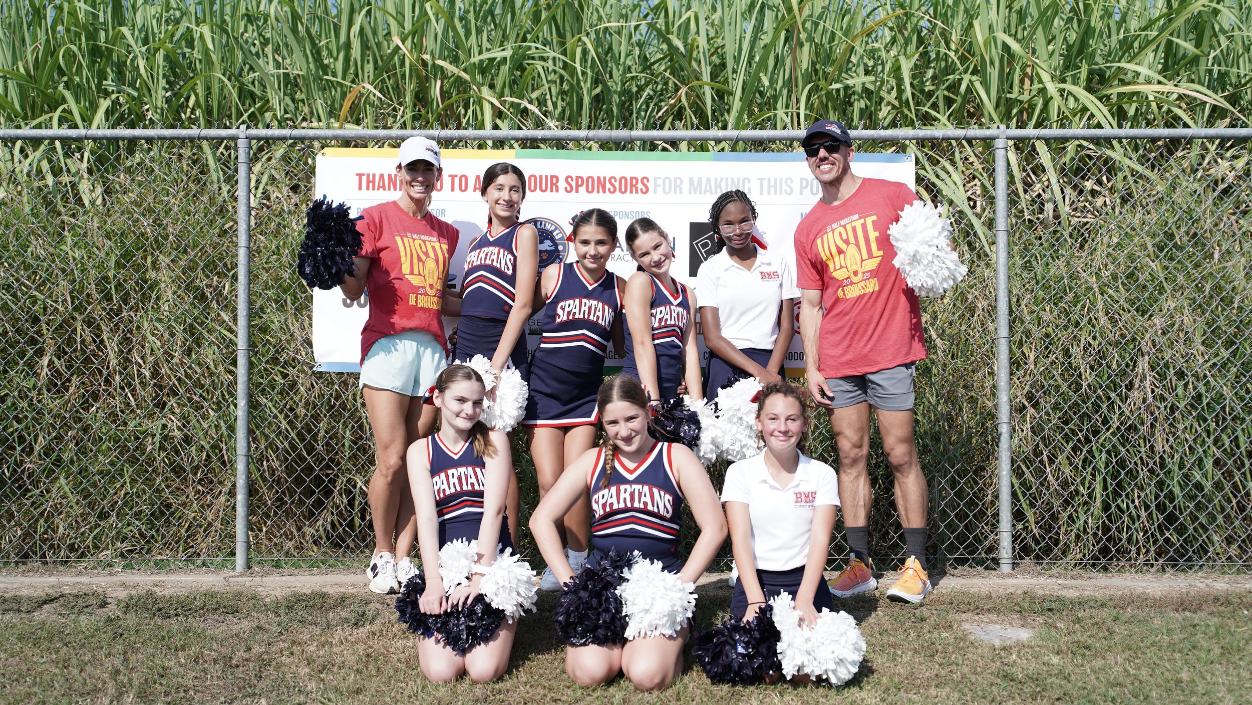 Group of children and adults posing in front of a sign at an outdoor event, some children are dressed as cheerleaders with pom-poms, and two adults are wearing red T-shirts related to a visit event.