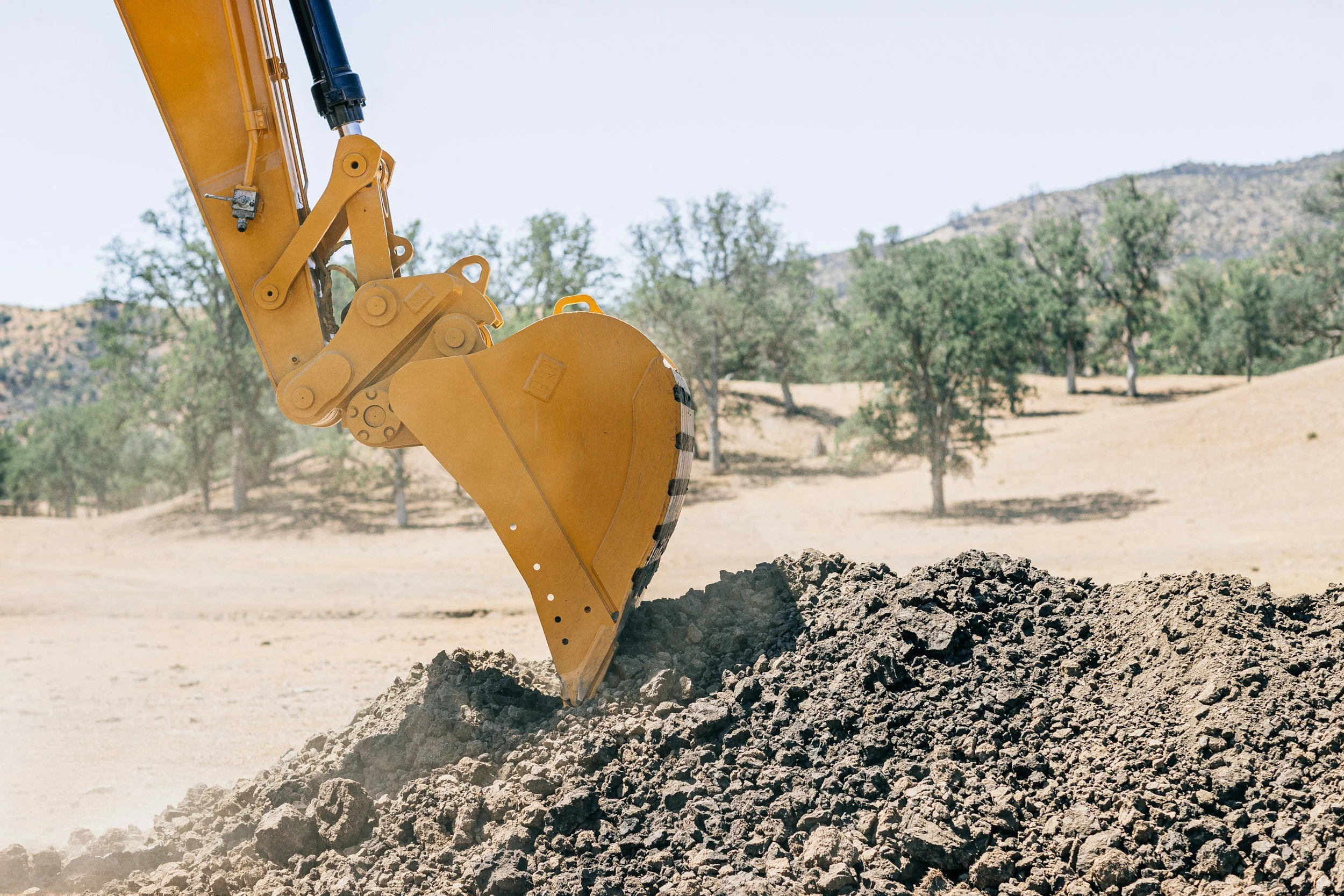 Close-up of a yellow excavator digging into a pile of dirt in a rural area with trees and hills in the background.