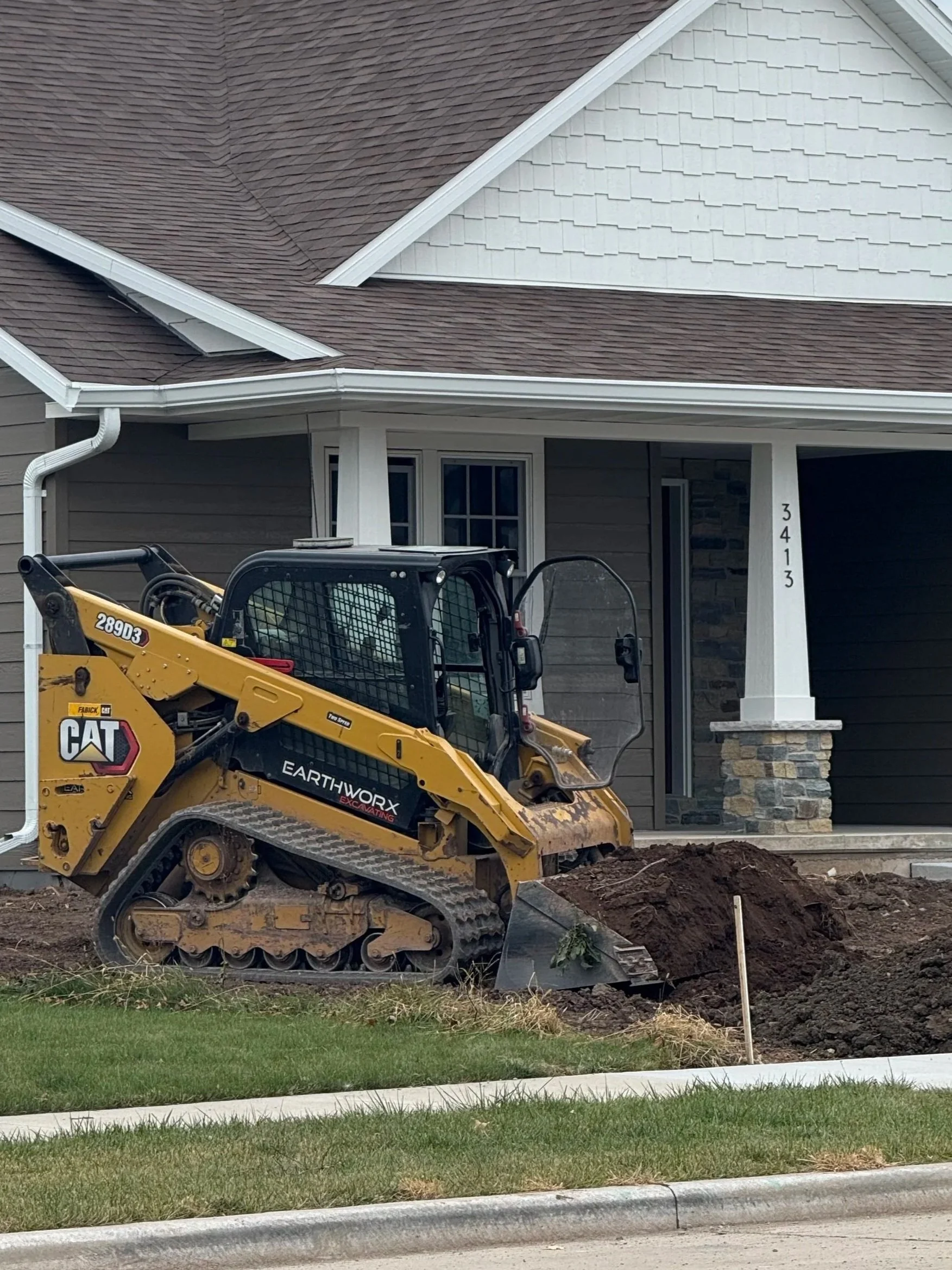 Construction vehicle working in front of a house with brown and white siding. The vehicle is a small yellow tracked excavator labeled 'CAT' and 'Earthworx Excavating,' digging a hole near the house's front yard. The house has a triangular roof with brown shingles and white trim, with the address 3413 visible on a white pillar.