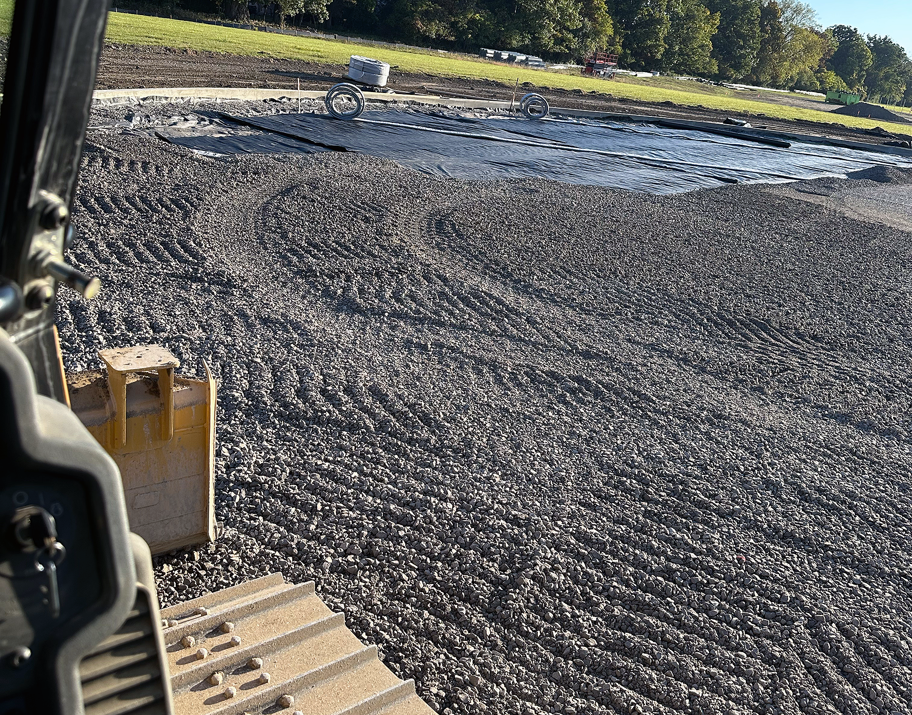 View from construction equipment overlooking a gravel-covered area with black plastic sheeting, surrounded by a grassy field and trees in the background.