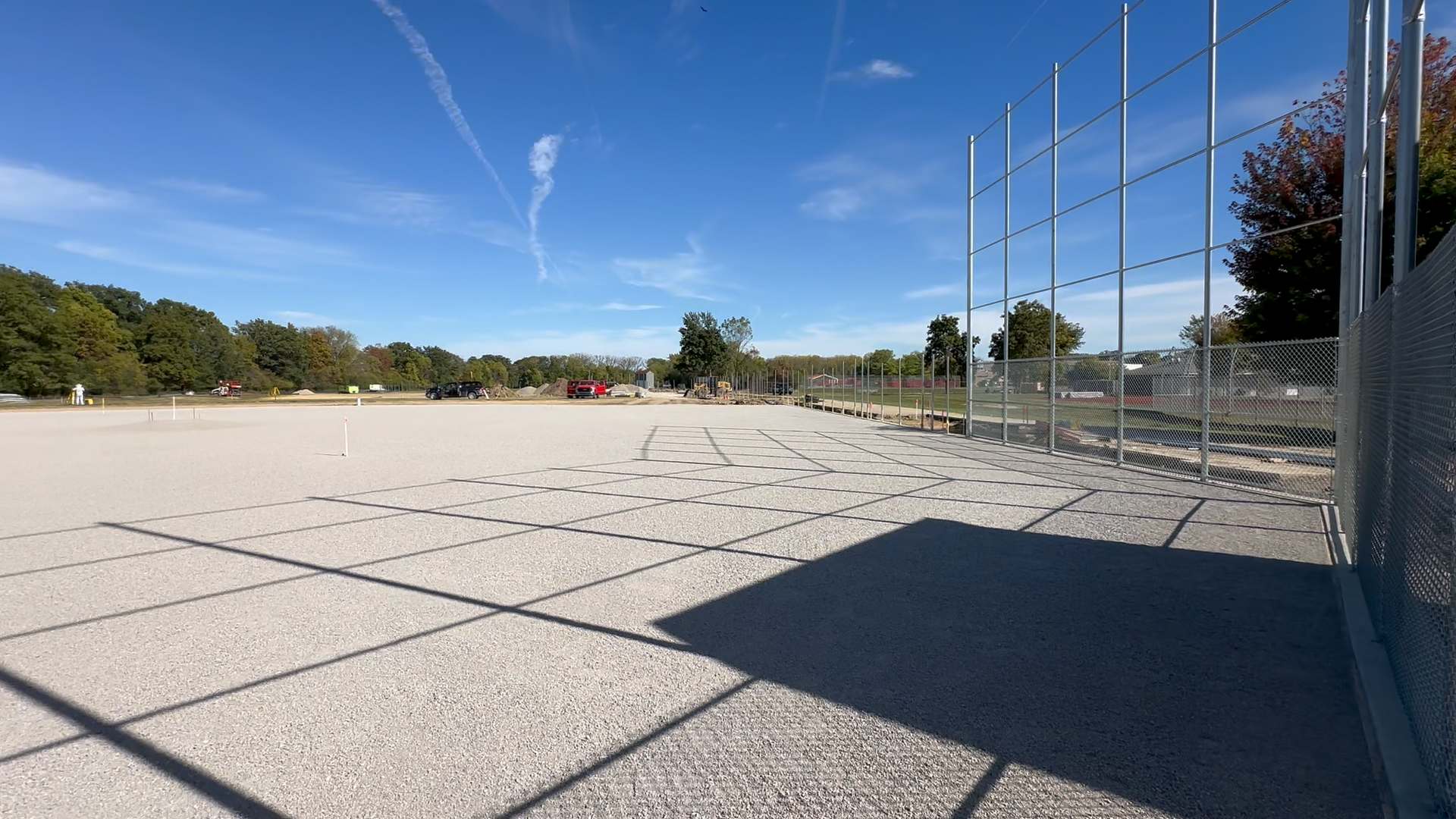 Finished earthwork construction project by Simone Company. A baseball field under final construction, with a gravel infield, a chain-link fence on the right, and construction vehicles in the background.