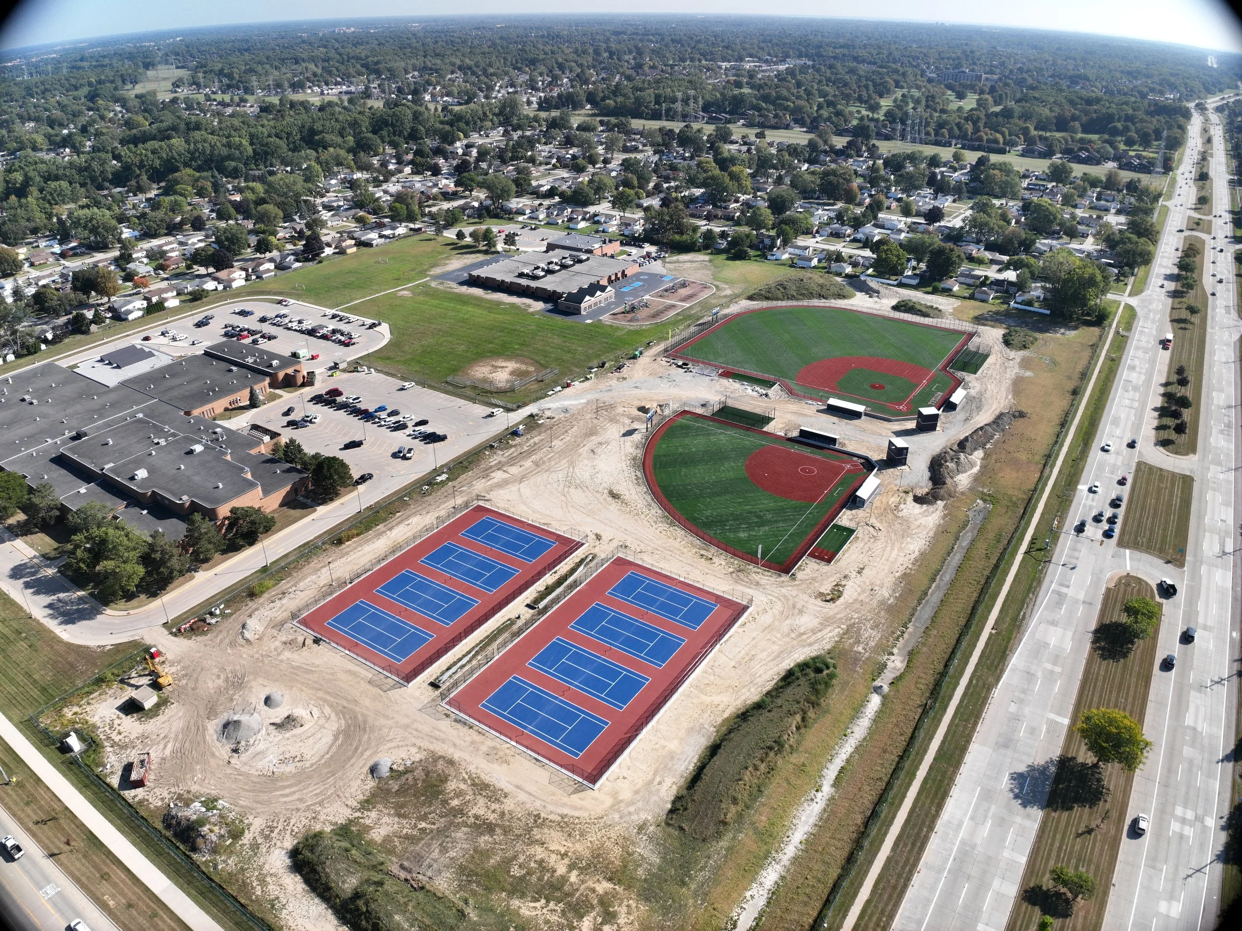 Aerial view of a sports complex under construction featuring multiple baseball fields, tennis courts, a parking lot, and nearby roads and residential areas.