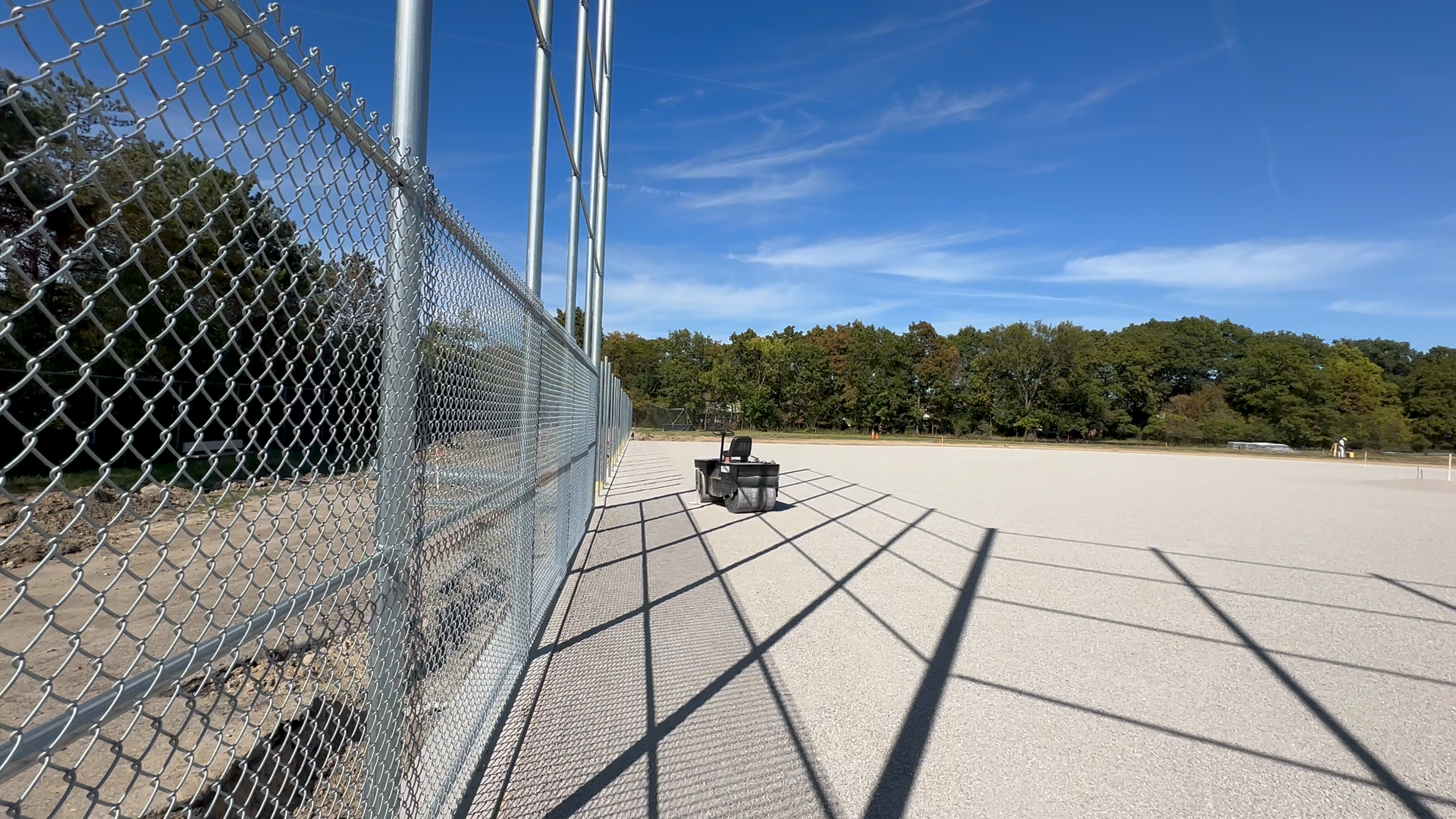 An empty sports field enclosed by a chain-link fence with shadows cast on the ground, a bench in the distance, and trees in the background under a blue sky with wispy clouds.