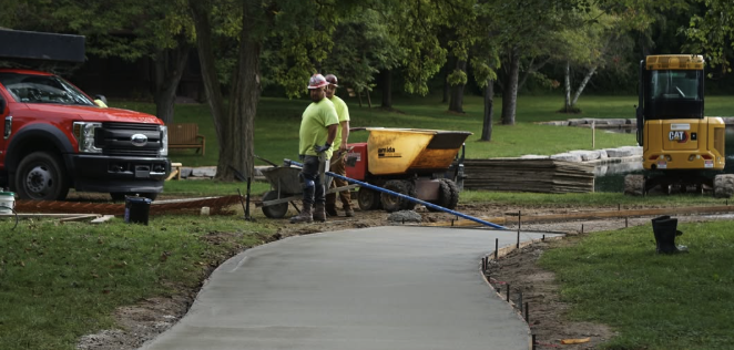 Two construction workers in neon yellow shirts and helmets work on a concrete pathway in a park, with a wheelbarrow nearby, a red pickup truck, and a yellow construction vehicle in the background.