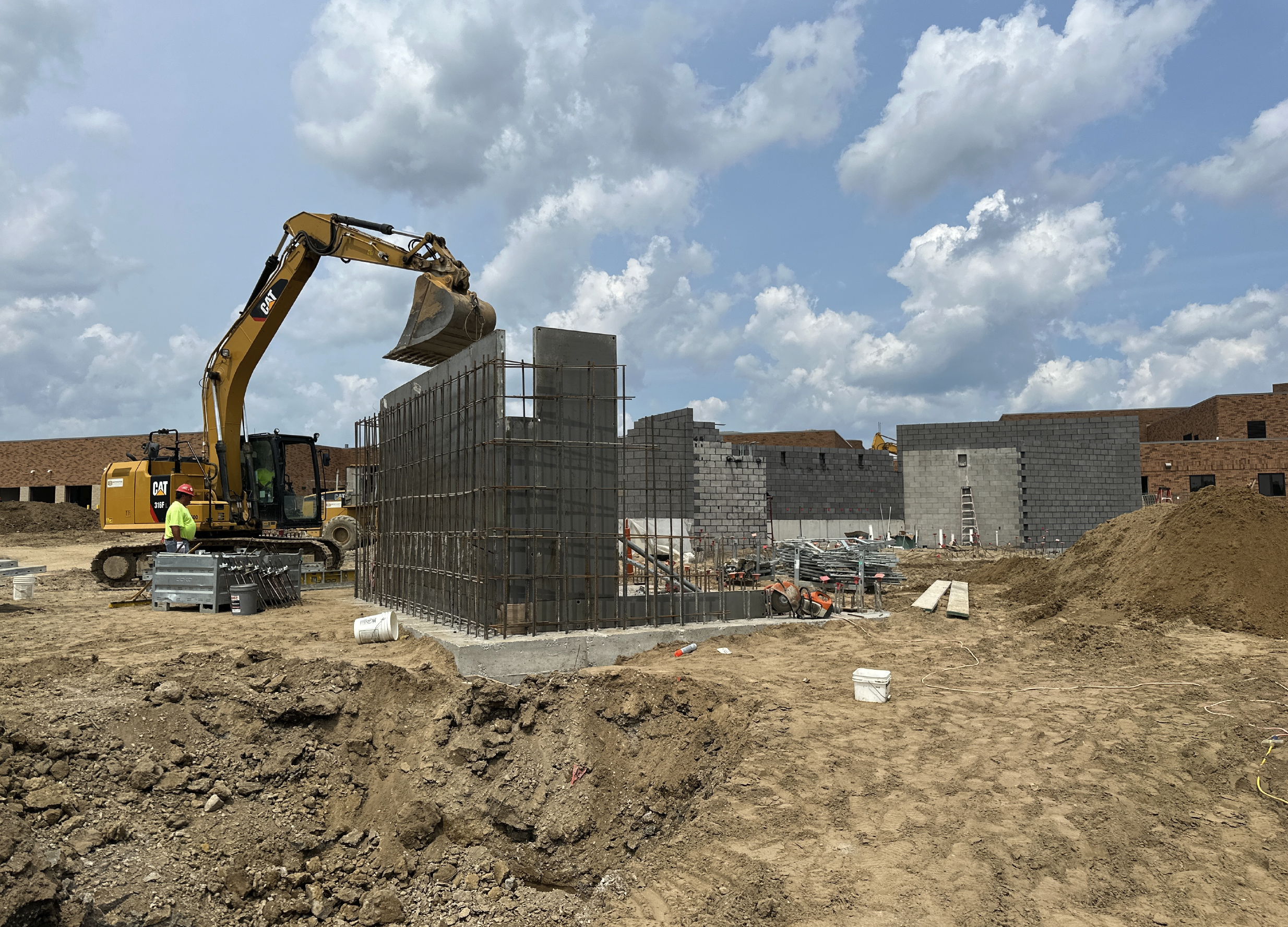 Construction site with a yellow excavator lifting a concrete wall section, and workers in safety gear. Scaffolding surrounds the construction of concrete walls with rebarb. The background shows partially built brick walls and a partly cloudy sky.
