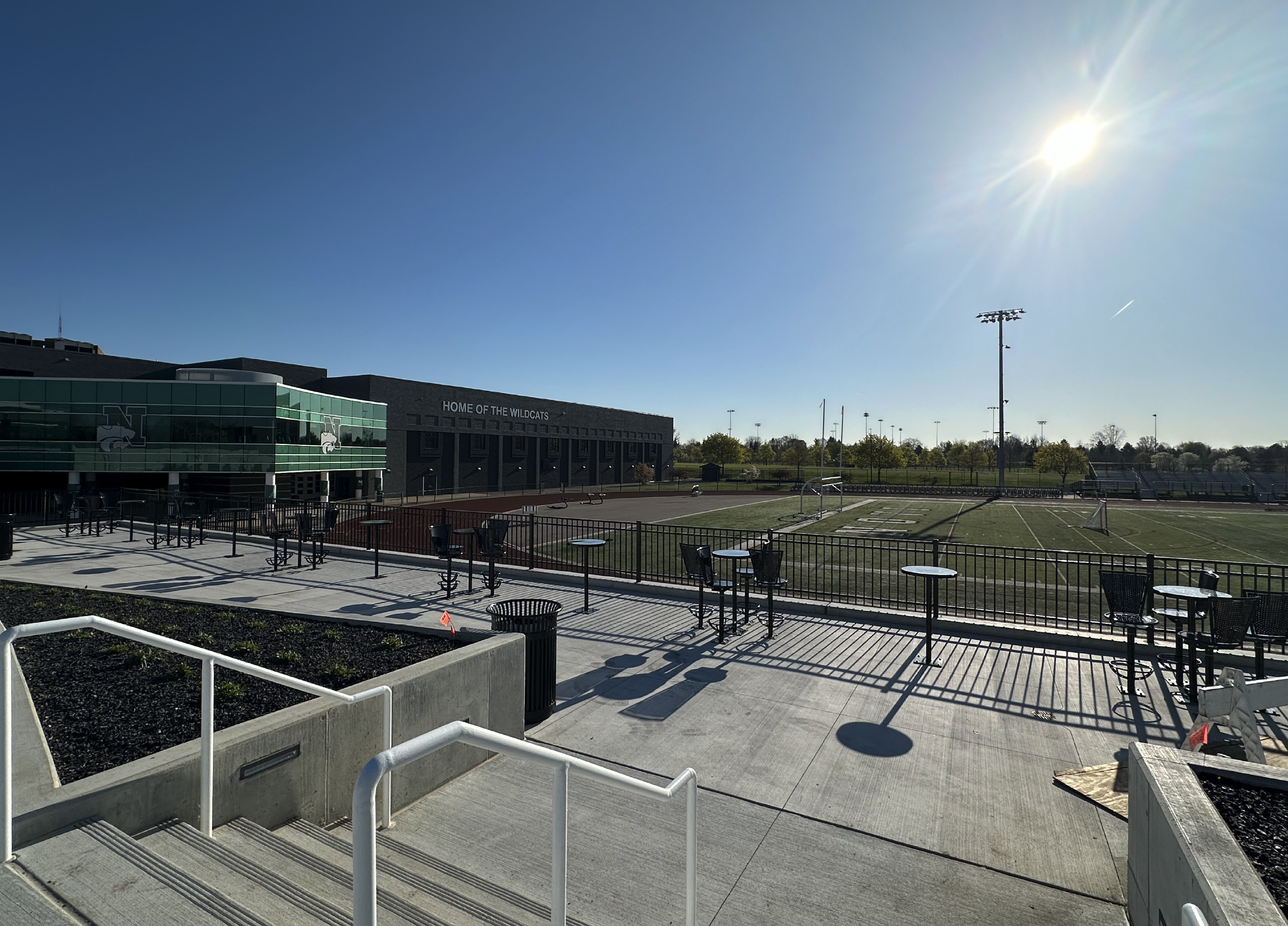 An empty outdoor sports stadium with a grass field, bleachers, and a building labeled 'Home of the Wildcats,' under a clear blue sky with the sun shining brightly.