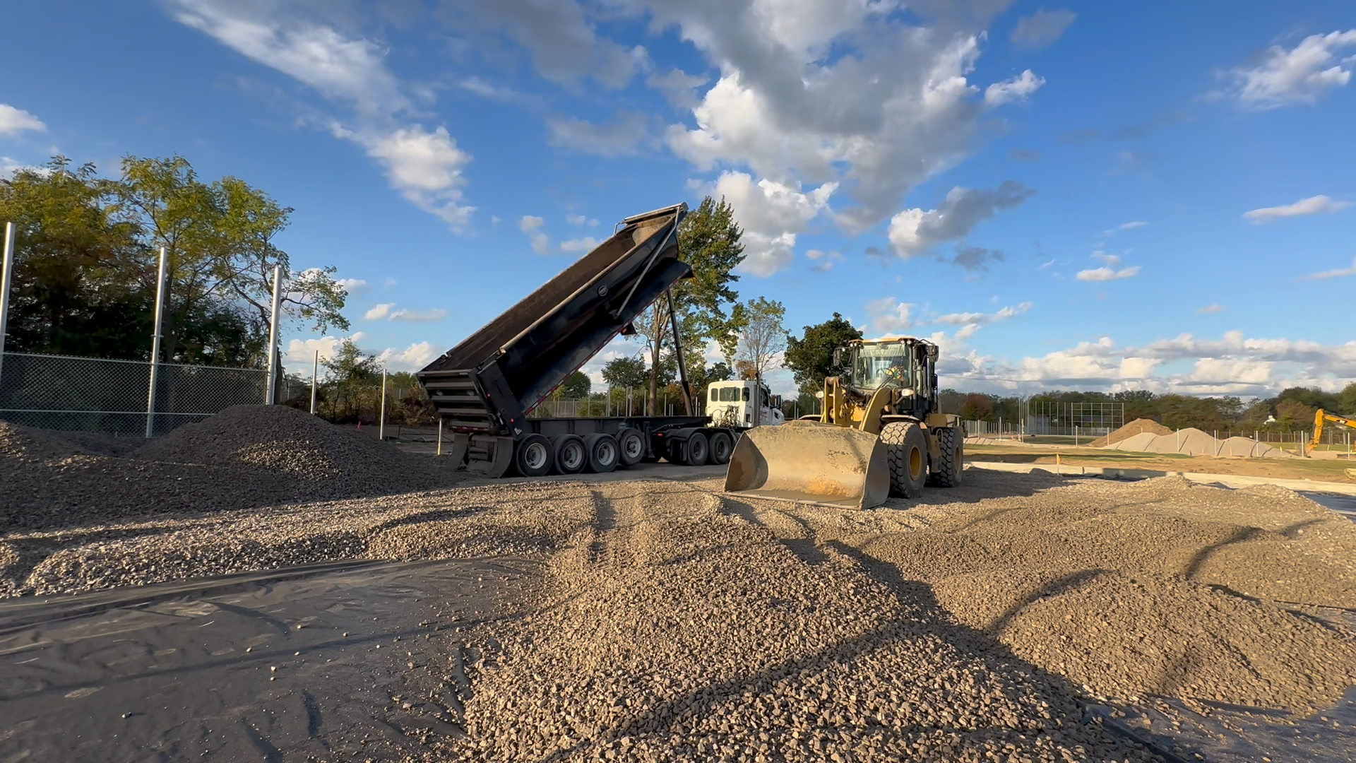 Earthwork construction site with a dump truck unloading gravel for a sports field build, a large front end loader moving gravel, dirt piles, construction fencing, and a blue sky with clouds.