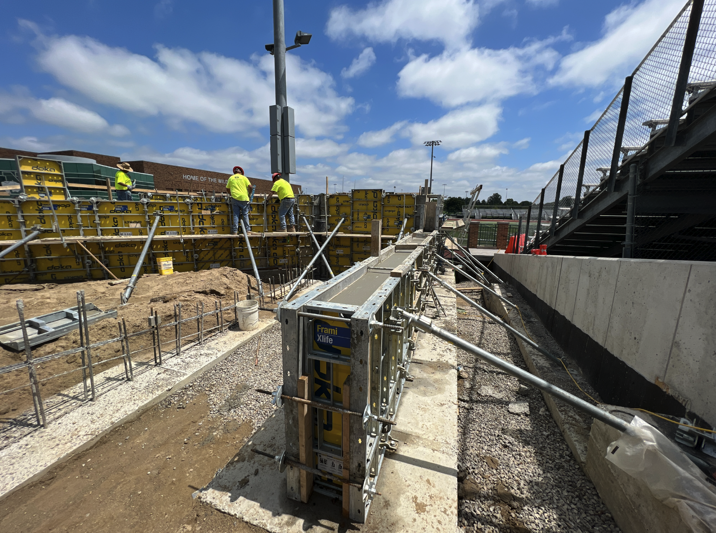 Construction workers in neon yellow safety shirts and red helmets working on a stadium construction site, with scaffolding and concrete walls under a partly cloudy sky.