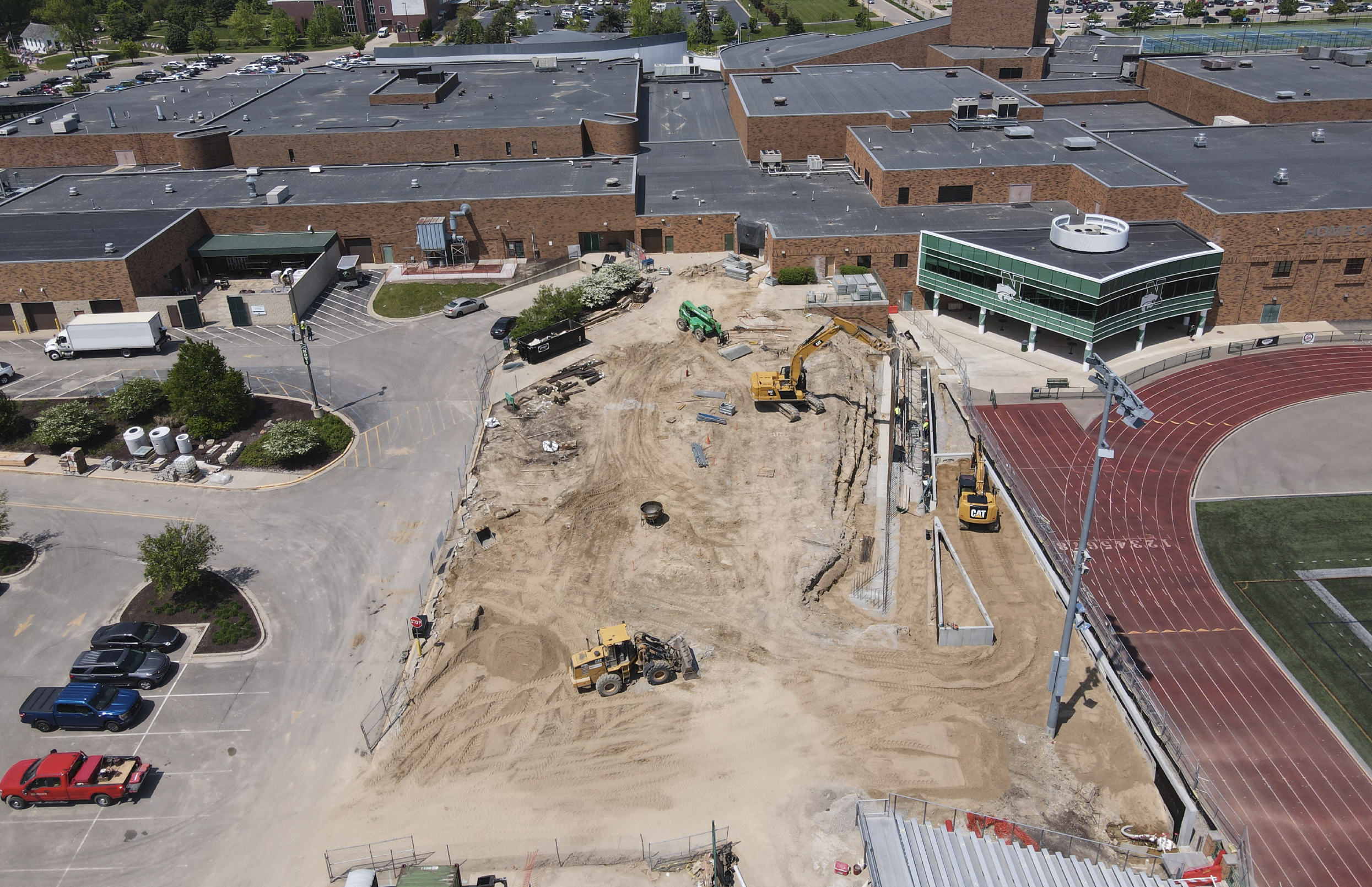 Aerial view of a construction site adjacent to a school athletic track and field. The site has heavy machinery and dirt, with no completed structures yet. Nearby, there are parked cars, a school building with a green and white facade, and a running track.