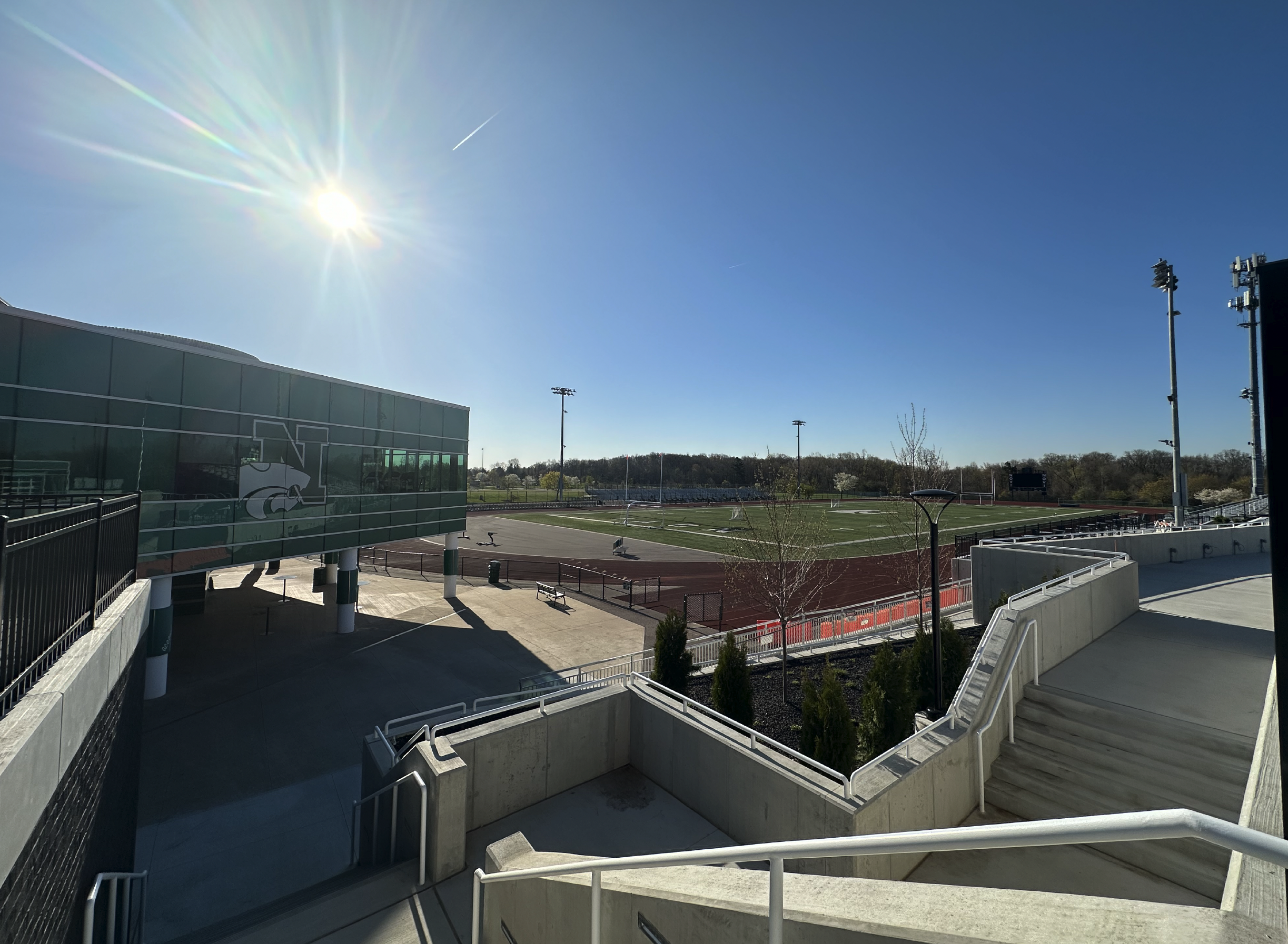 Empty football field with bright sunlight, clear blue sky, and surrounding athletic facilities.