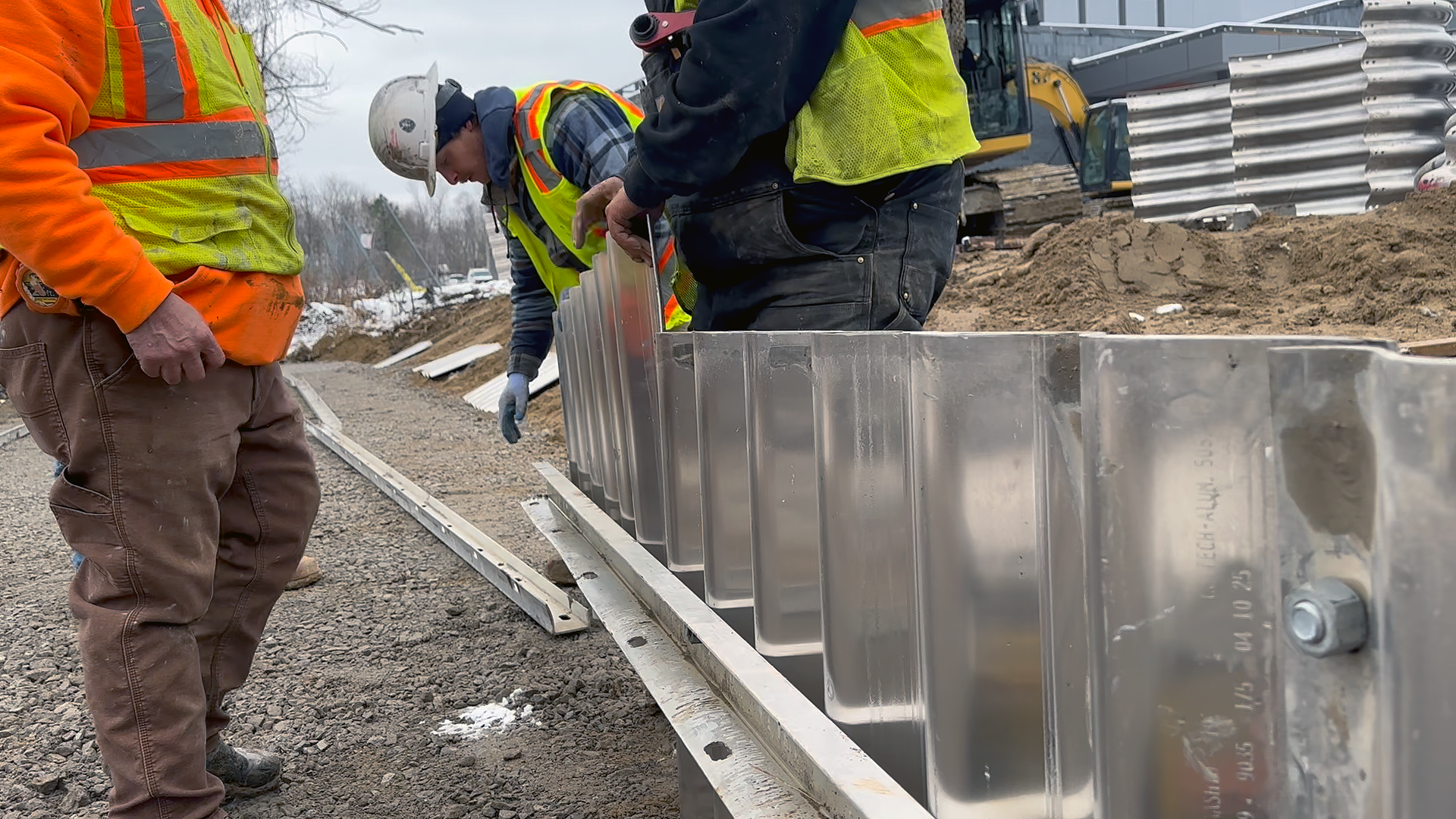 Underground utility construction workers in safety vests and helmets installing a metal barrier for underground drainage on a compacted gravel surface at a construction site with an excavator in the background.