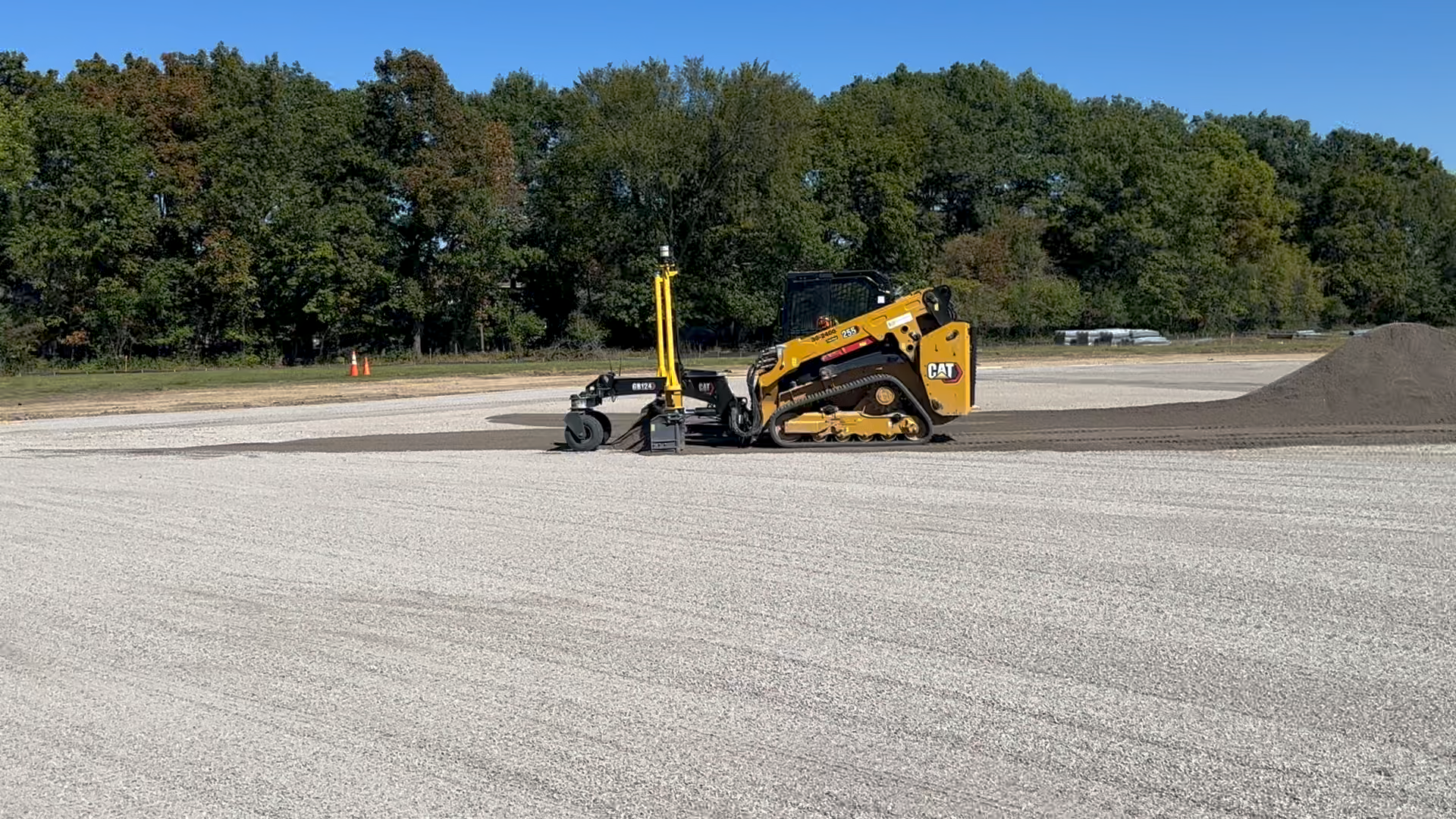 A yellow CAT compact construction vehicle working on a gravel surface with a small pile of gravel nearby and trees in the background under a clear blue sky.