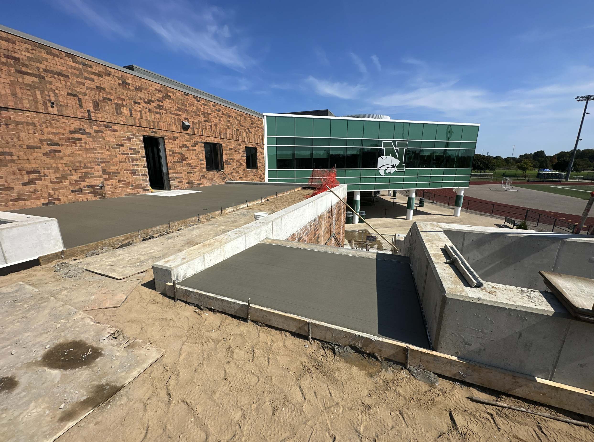 Construction site of a school building with fresh concrete slabs, brick walls, and a modern glass extension featuring a large green logo of a Spartan helmet and the letter 'N'. A running track and athletic fields are visible in the background under a clear blue sky.