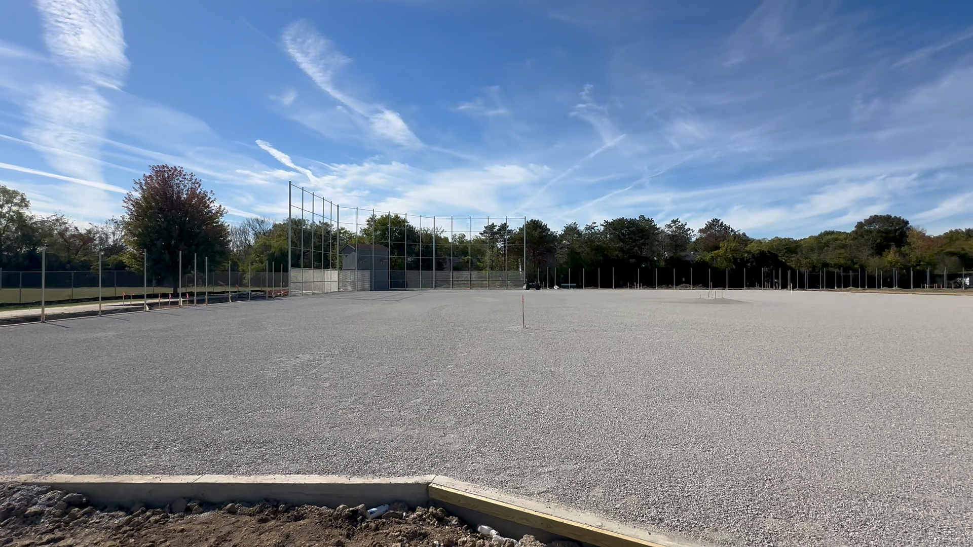 Finished earthwork construction project by Simone Companies. A finished empty baseball field with a gravel infield prepared for artificial turf application, a backstop with tall fence, and a tree-lined background under a partly cloudy sky.