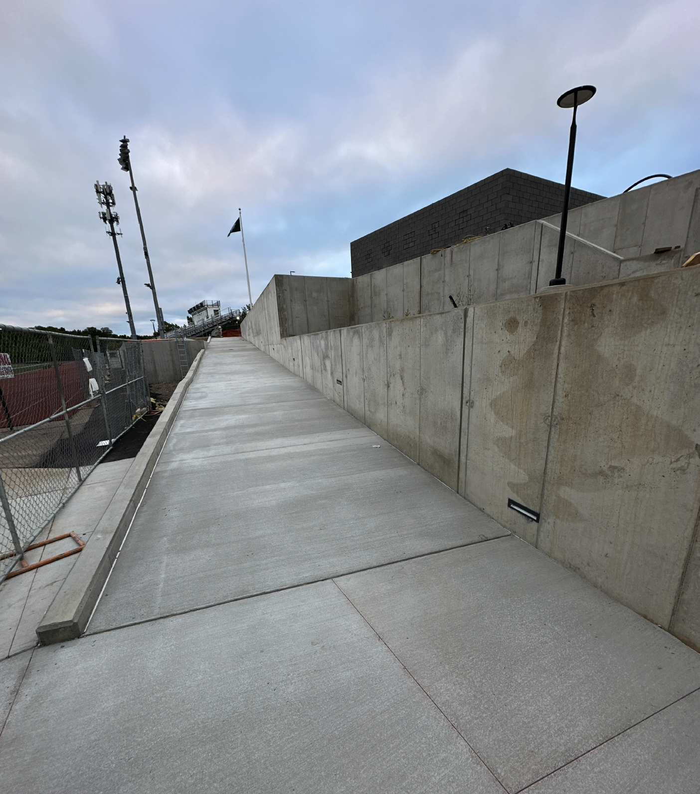 Concrete walkway with a sloped incline, concrete wall on the right side, construction fencing on the left, and a black building in the background. Overcast sky.